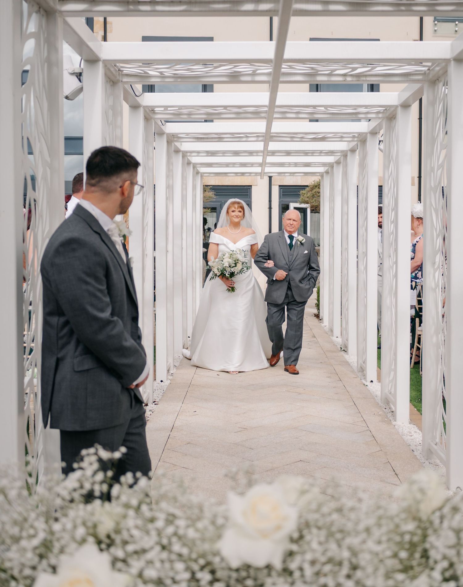 Wedding party walking through an elegant white pergola corridor decorated with flowers.