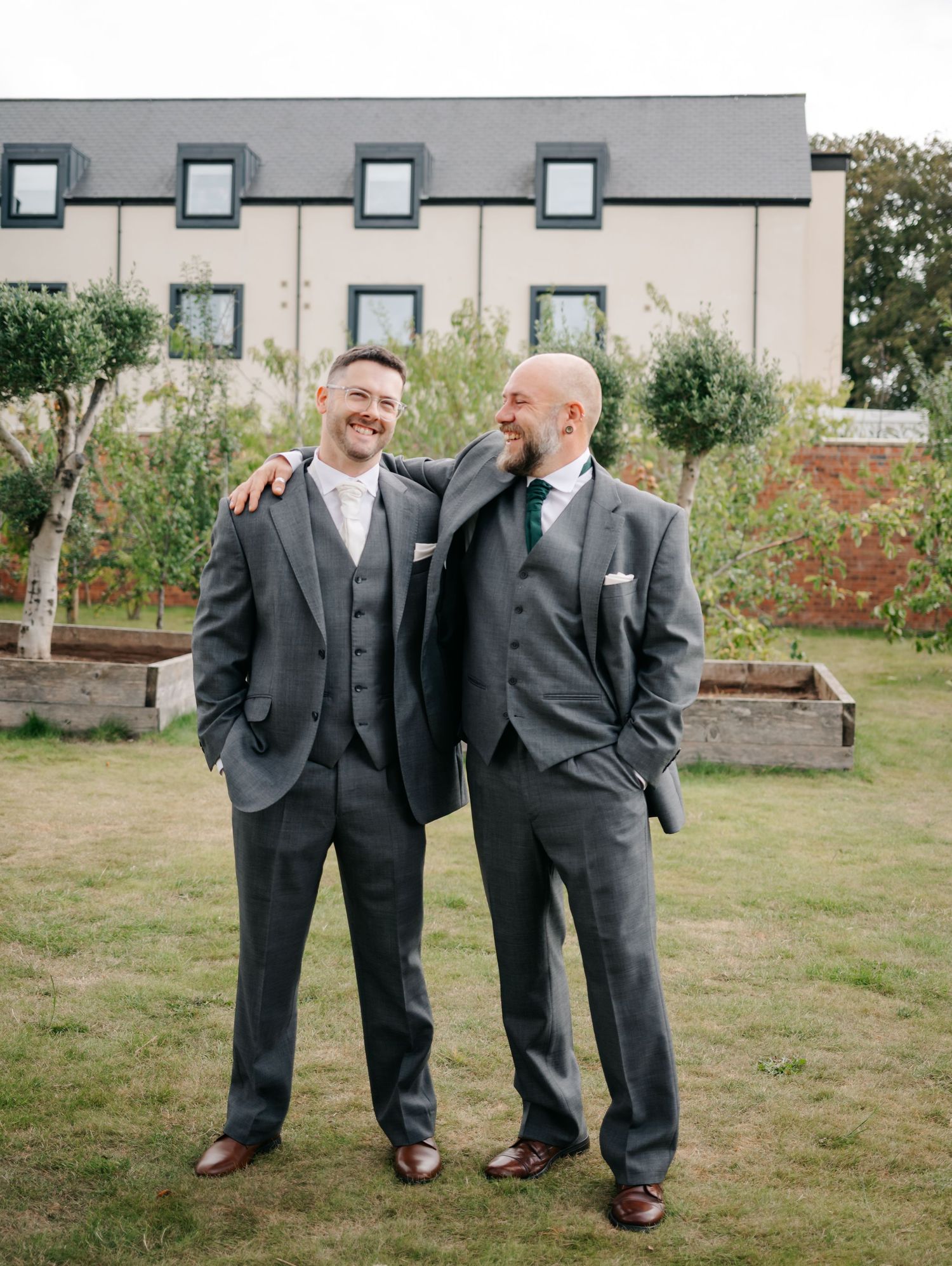 Two people in dark suits sharing a lighthearted moment on a grassy lawn.