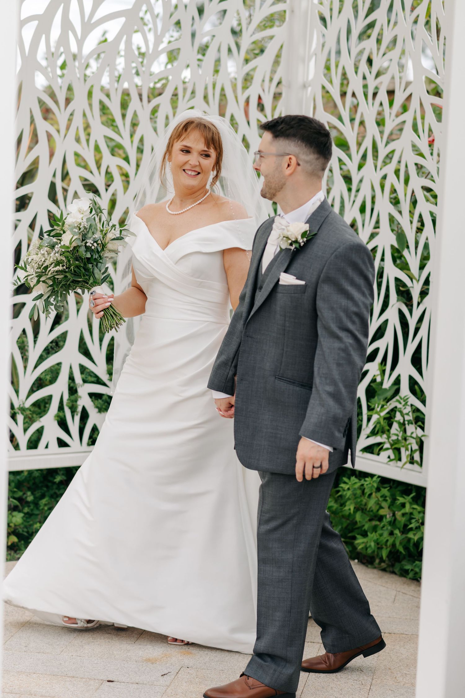 A wedding couple poses under an elegant white lattice archway with greenery in the background.