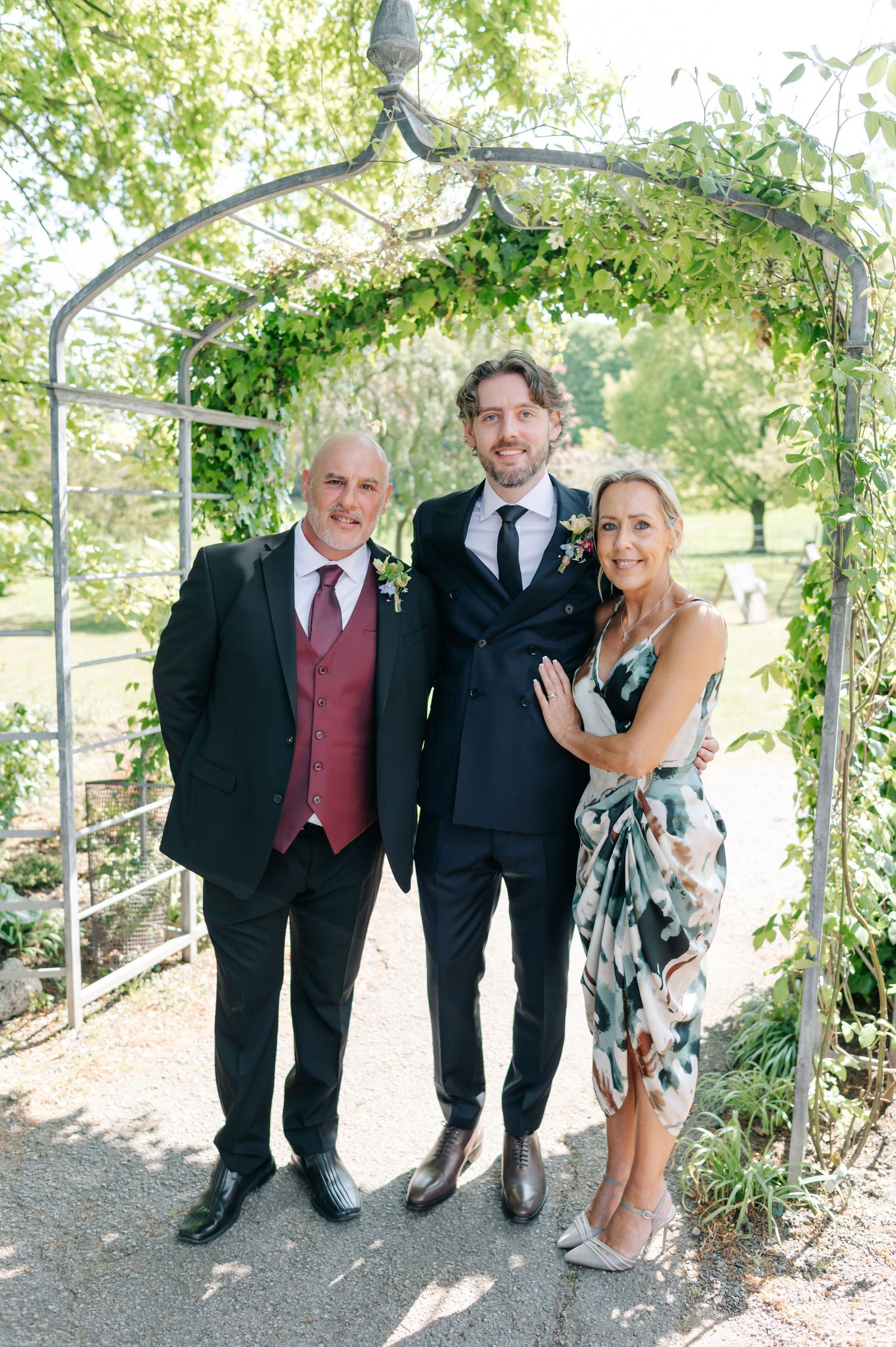 Three people pose together under an ivy-covered garden arch during an outdoor celebration.