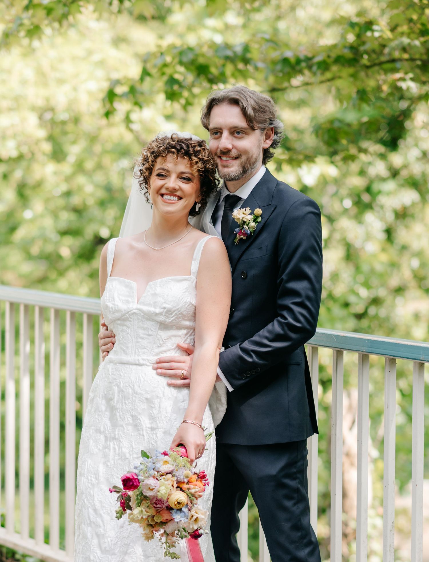Newlyweds pose together on outdoor balcony with floral bouquet and natural backdrop.