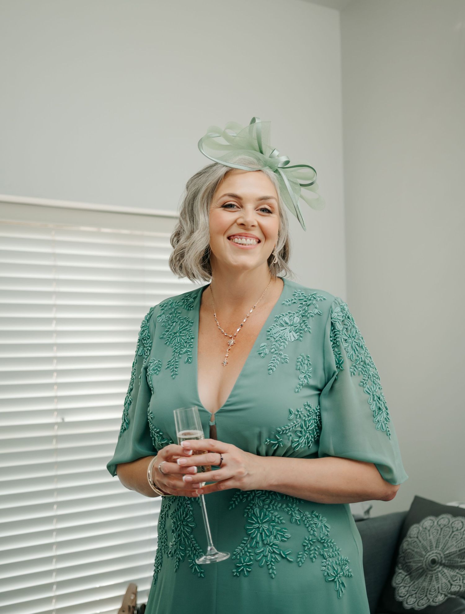 A guest in a mint green lace dress and fascinator holds a champagne glass at a wedding celebration.