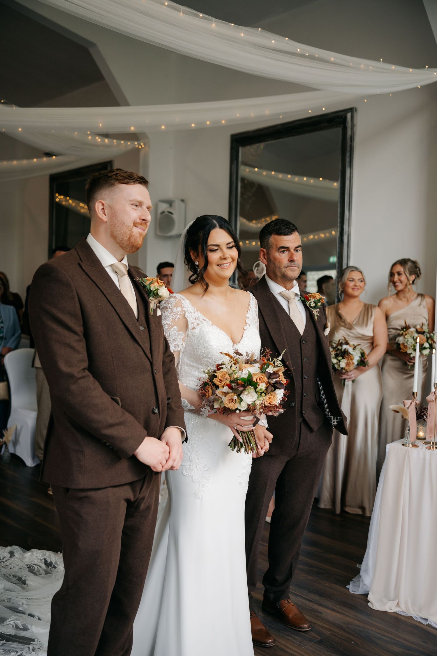 Wedding party members in brown suits and white dresses stand together during an indoor celebration with string lights.