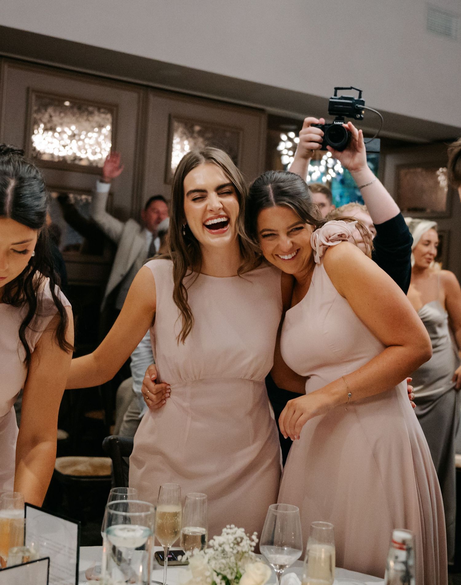 Bridesmaids in blush pink dresses share laughs during wedding reception.