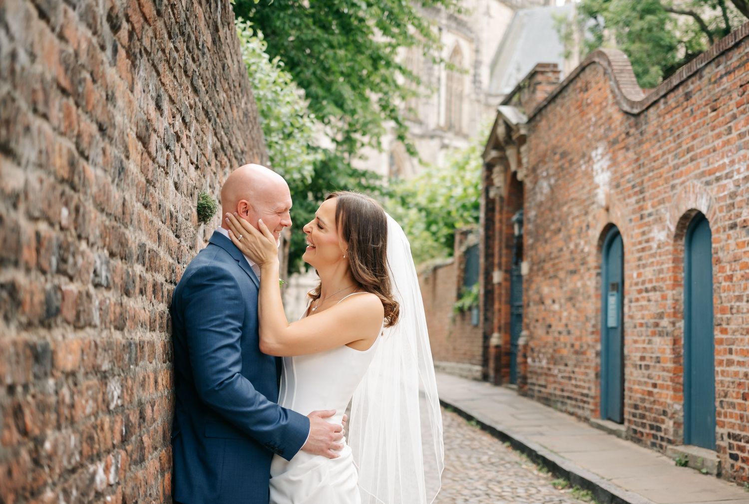 Bride and groom share a romantic moment leaning against a rustic brick wall on a cobblestone alley.