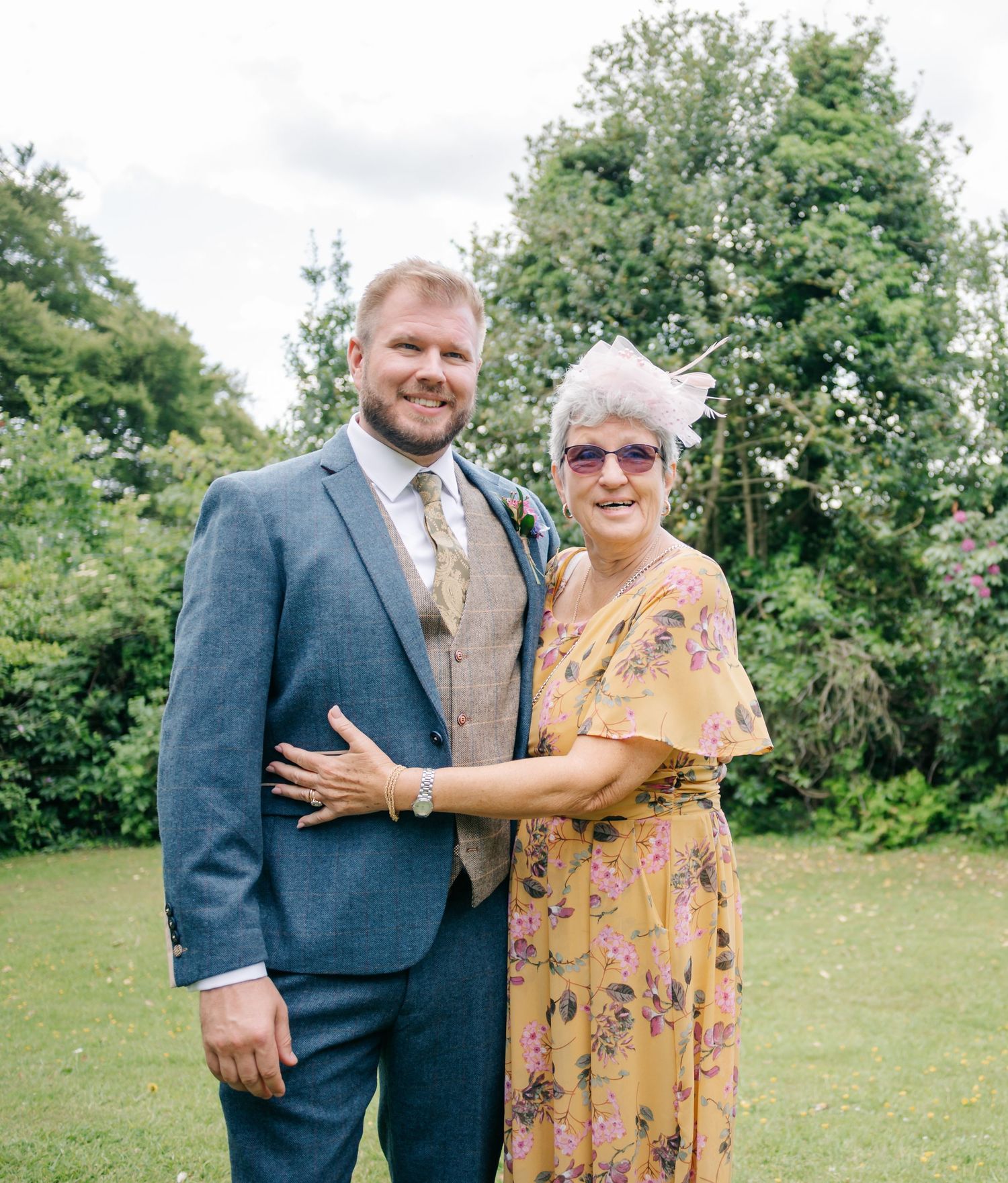 Elegant wedding guests pose together on a sunny lawn with green trees in the background.