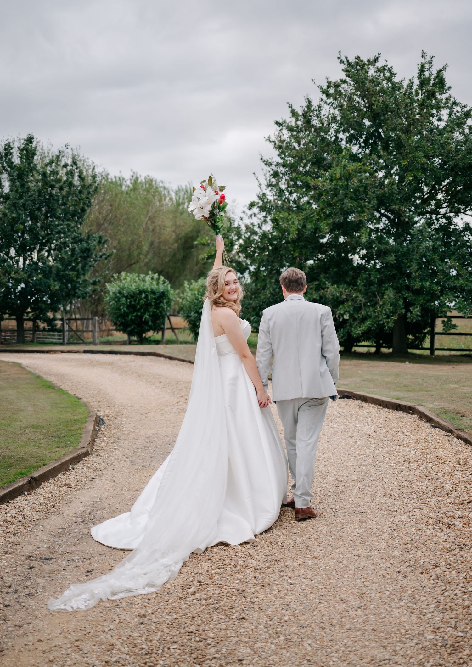 A couple walking on a gravel path surrounded by green trees during their outdoor wedding celebration.