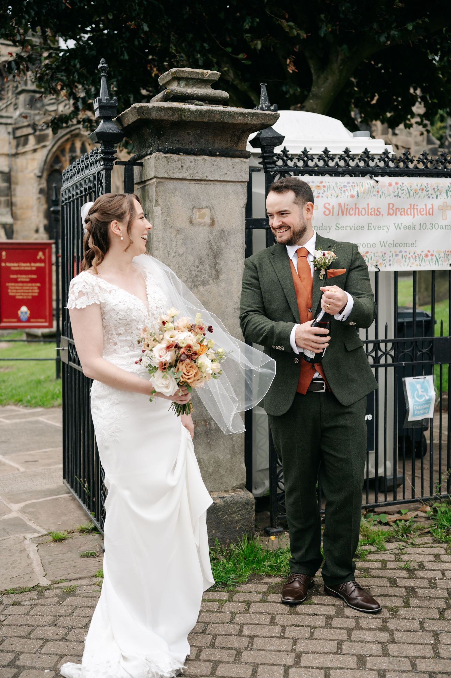 Laughing bride in a veil and fitted wedding dress shares a joyful moment with her groom outside the St Nicholas church gates.