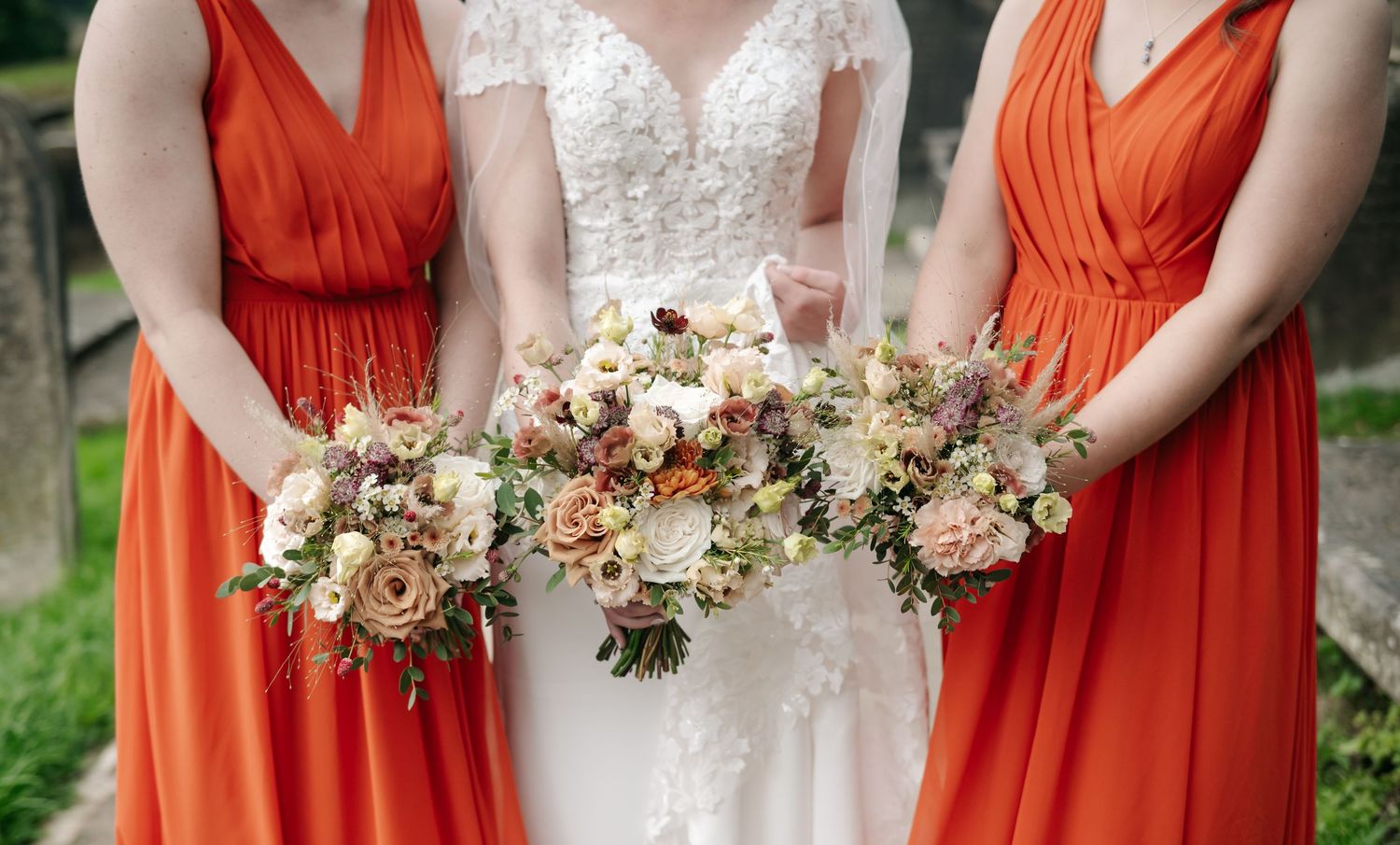 A bride in a lace wedding dress stands between two bridesmaids in orange dresses, all holding rustic bouquets of peach and cream flowers.