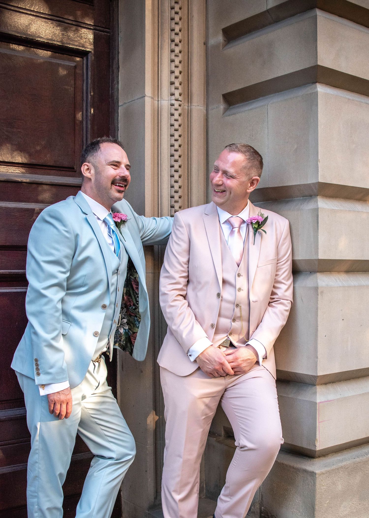 Two people in light colored suits laugh together while posing outside a historic building.