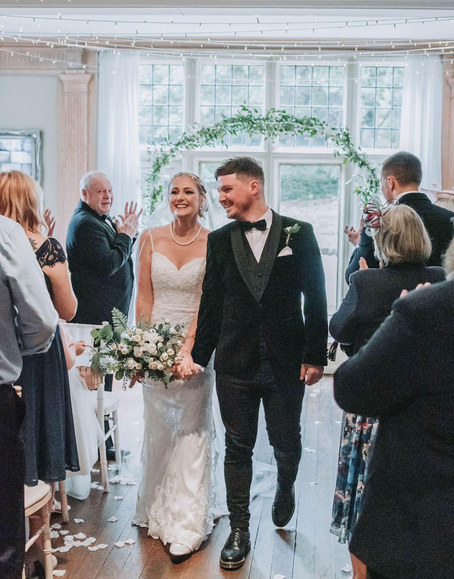 Newlyweds walk down aisle while guests celebrate in bright indoor venue with floral arch.