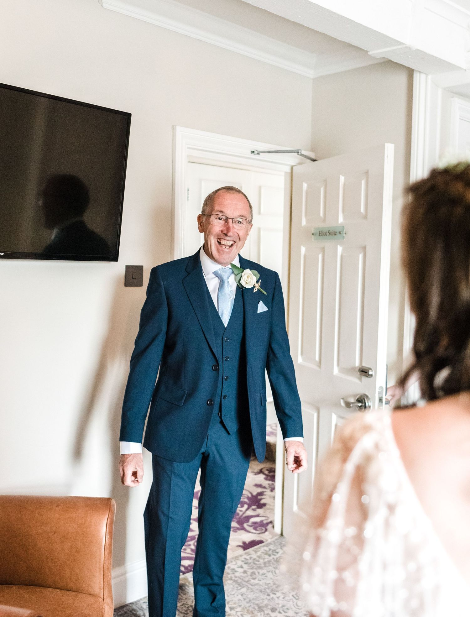 Someone in a navy three-piece suit with boutonniere smiles in reception room.