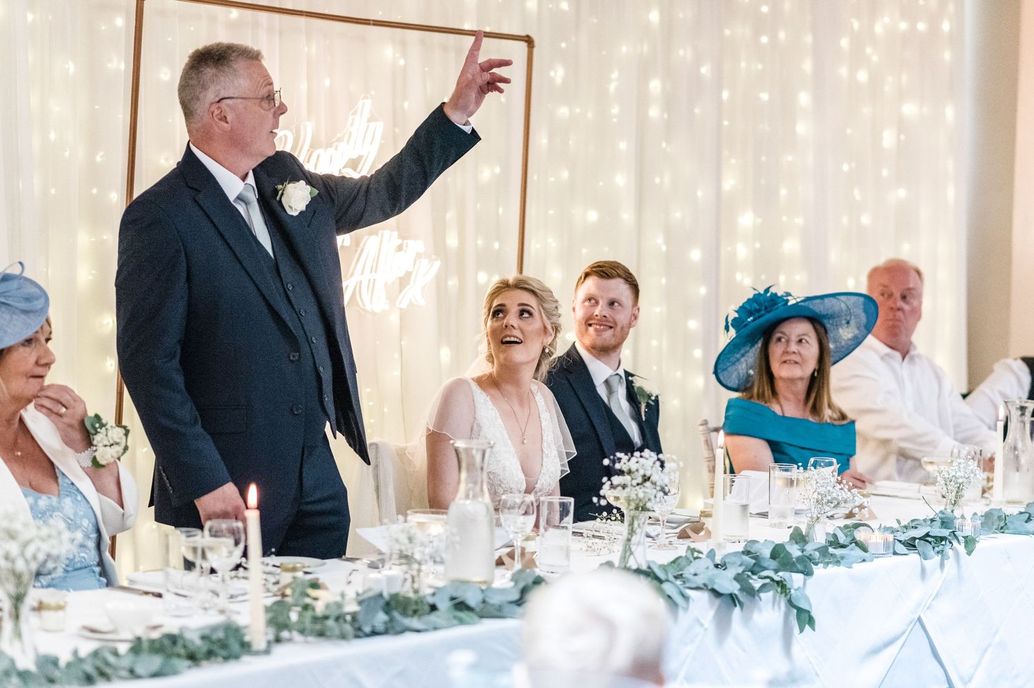 A reception speech being given at a wedding with guests seated at a decorated head table.