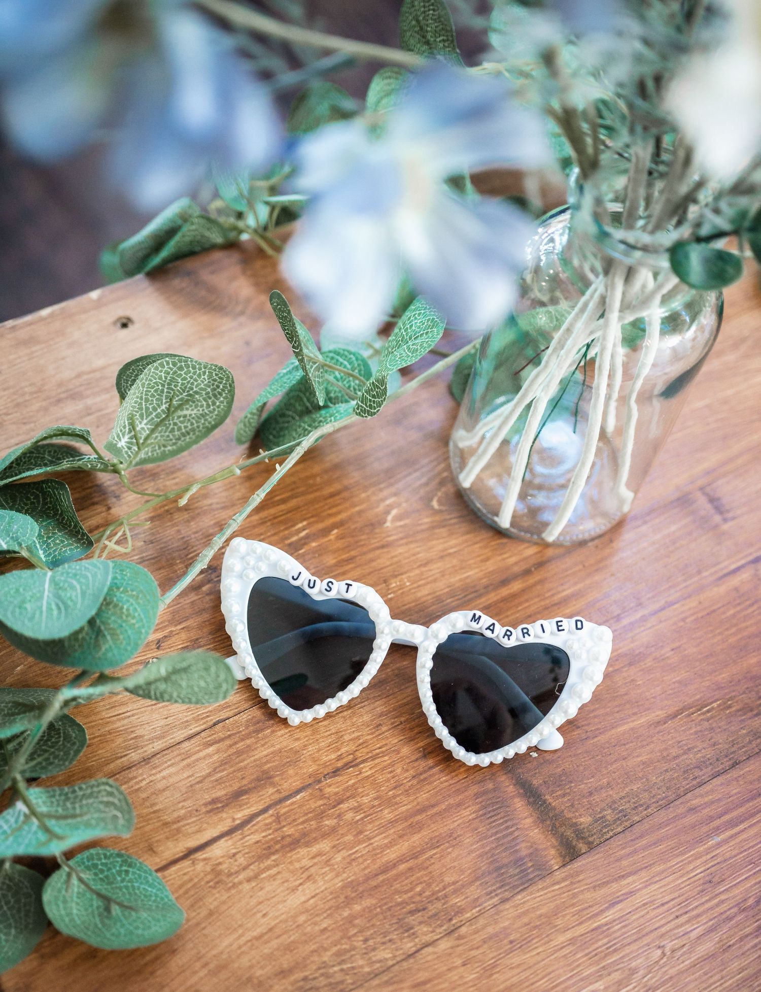 Heart-shaped white sunglasses displayed next to eucalyptus stems and white flowers on wooden surface.