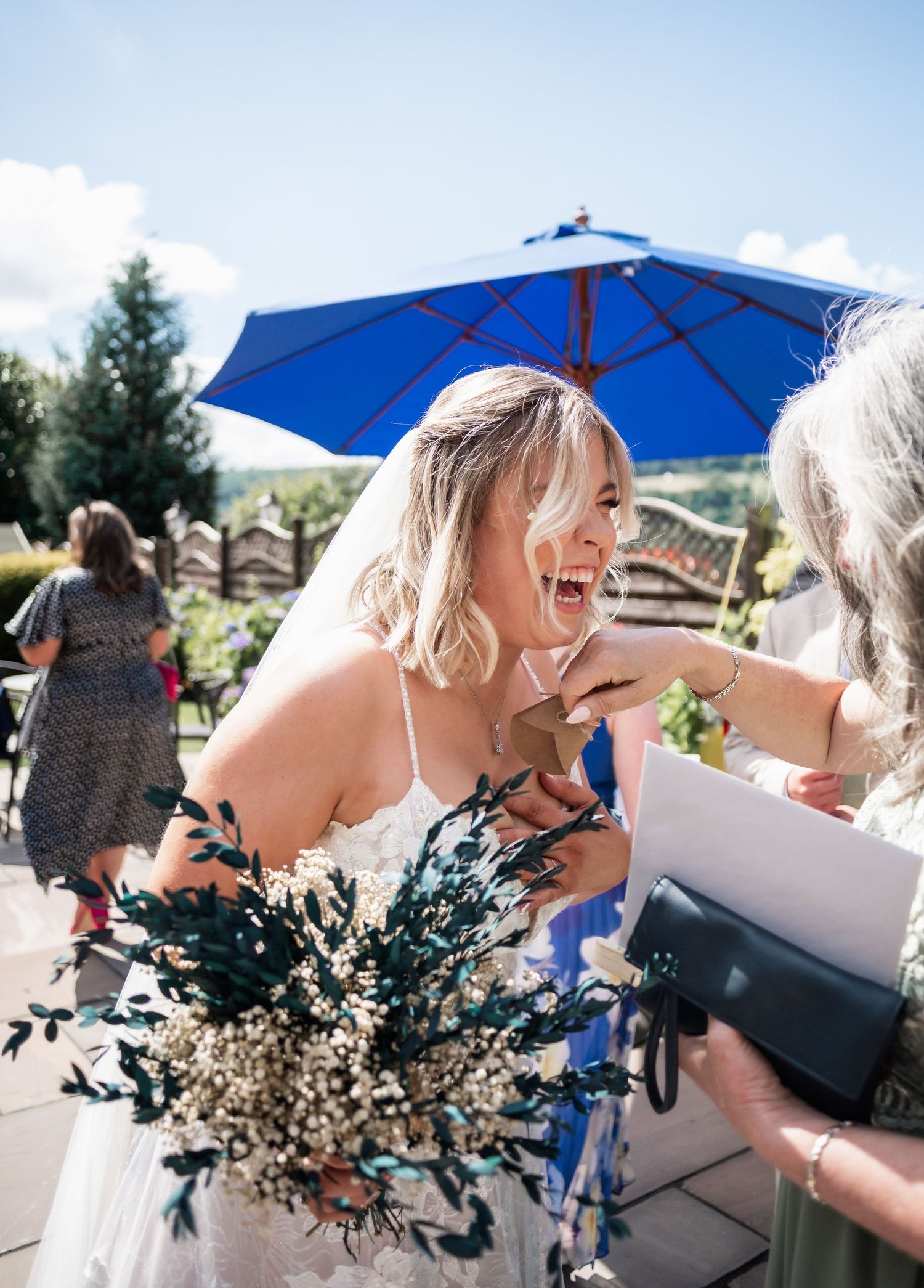 A joyful candid wedding moment under a bright blue umbrella with a rustic dried floral bouquet.