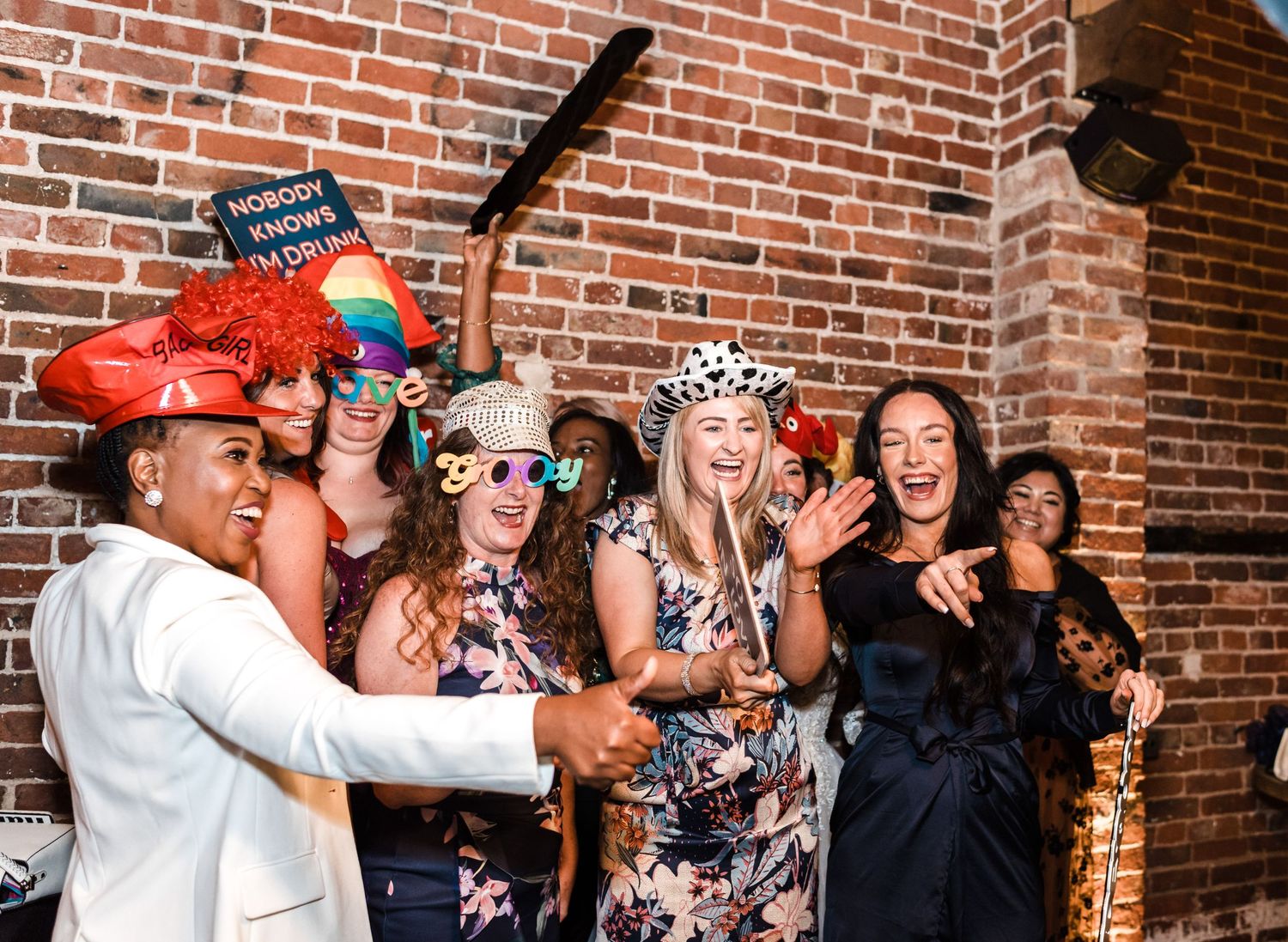 Group of wedding guests laugh while posing with photo booth props against brick wall.