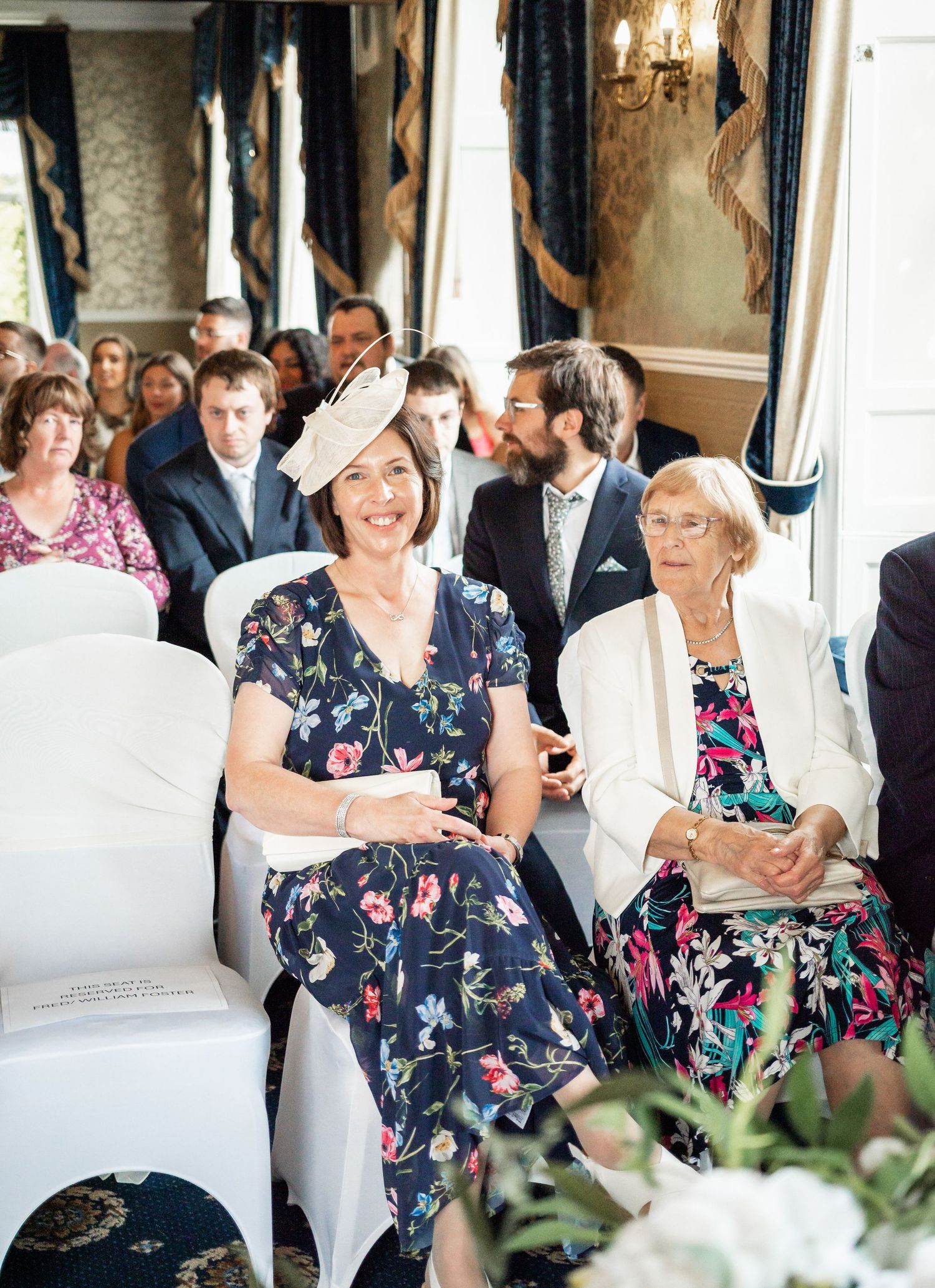 Wedding guests in floral dresses seated in elegant venue with white chairs and blue drapes.