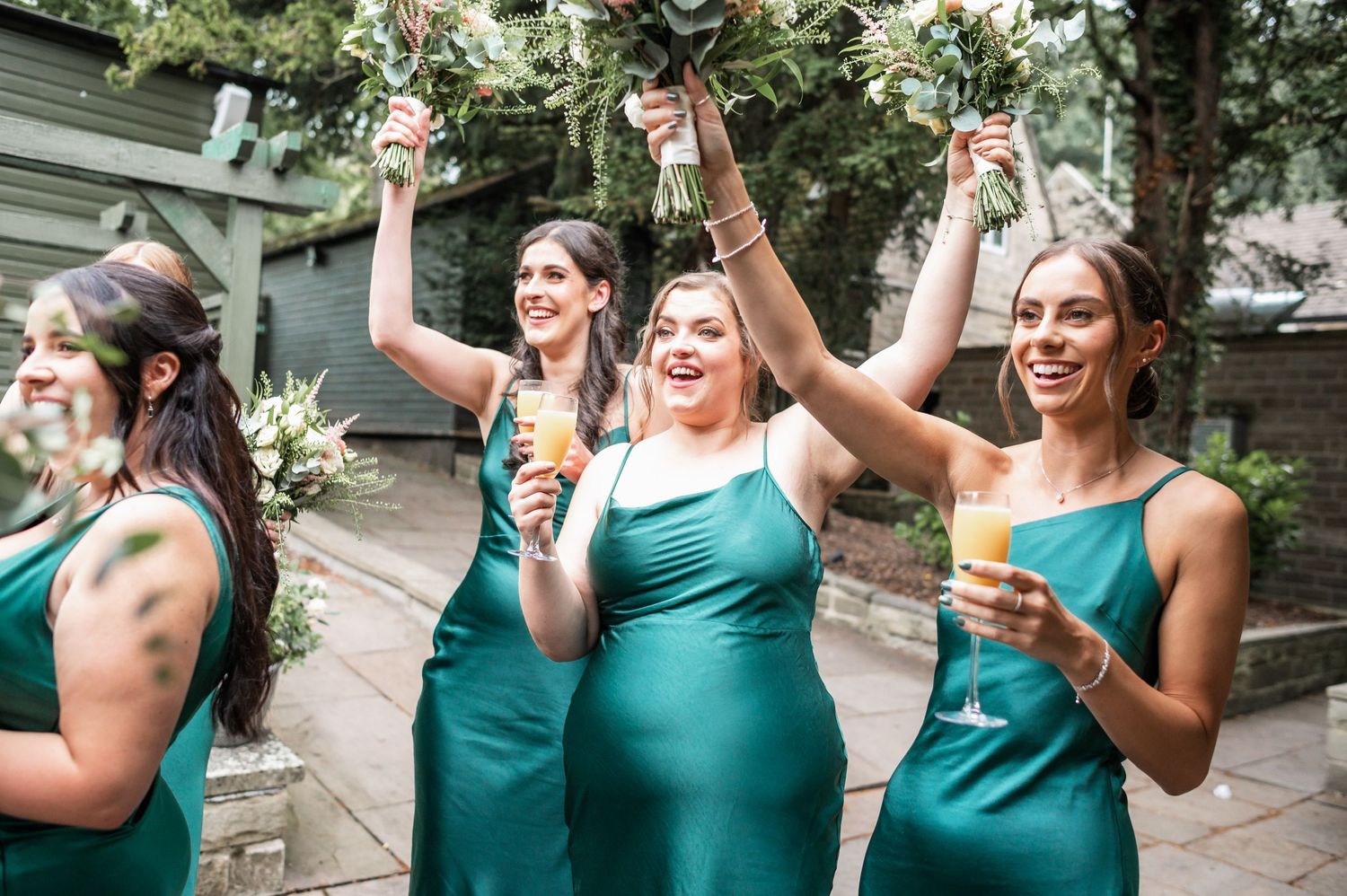 Bridesmaids in emerald green dresses celebrate with champagne glasses raised high.