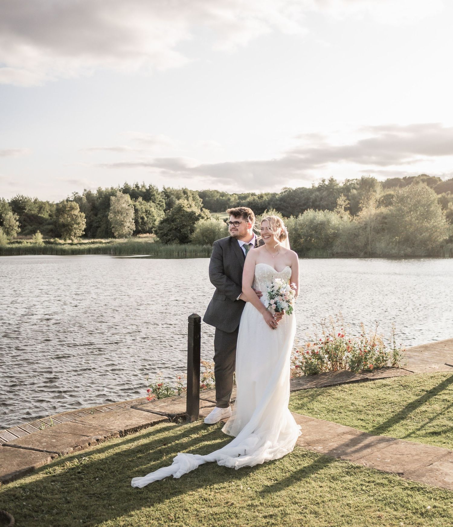 Romantic waterfront portrait of newlyweds standing together by lake at sunset.