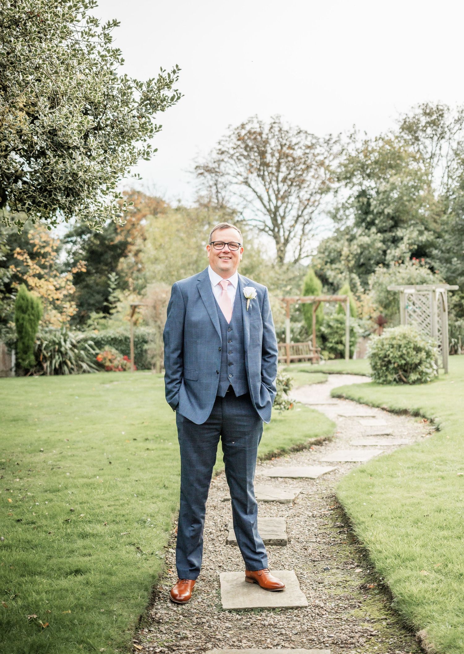 Person in navy three-piece suit standing on garden path surrounded by greenery.