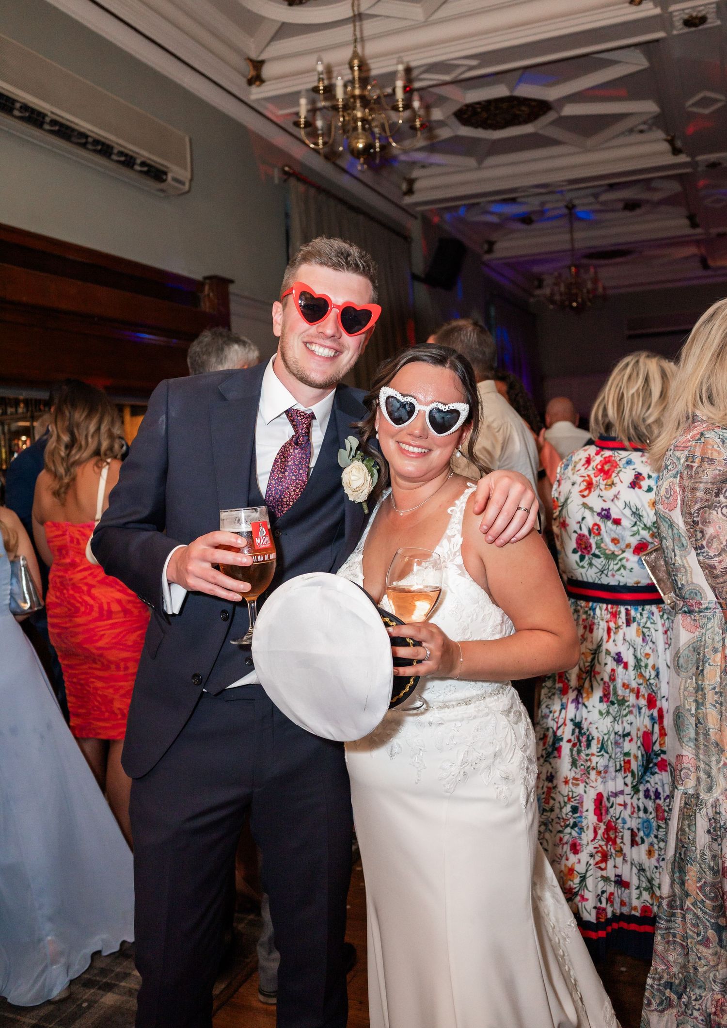 Couple poses playfully with drinks and heart sunglasses during wedding reception.