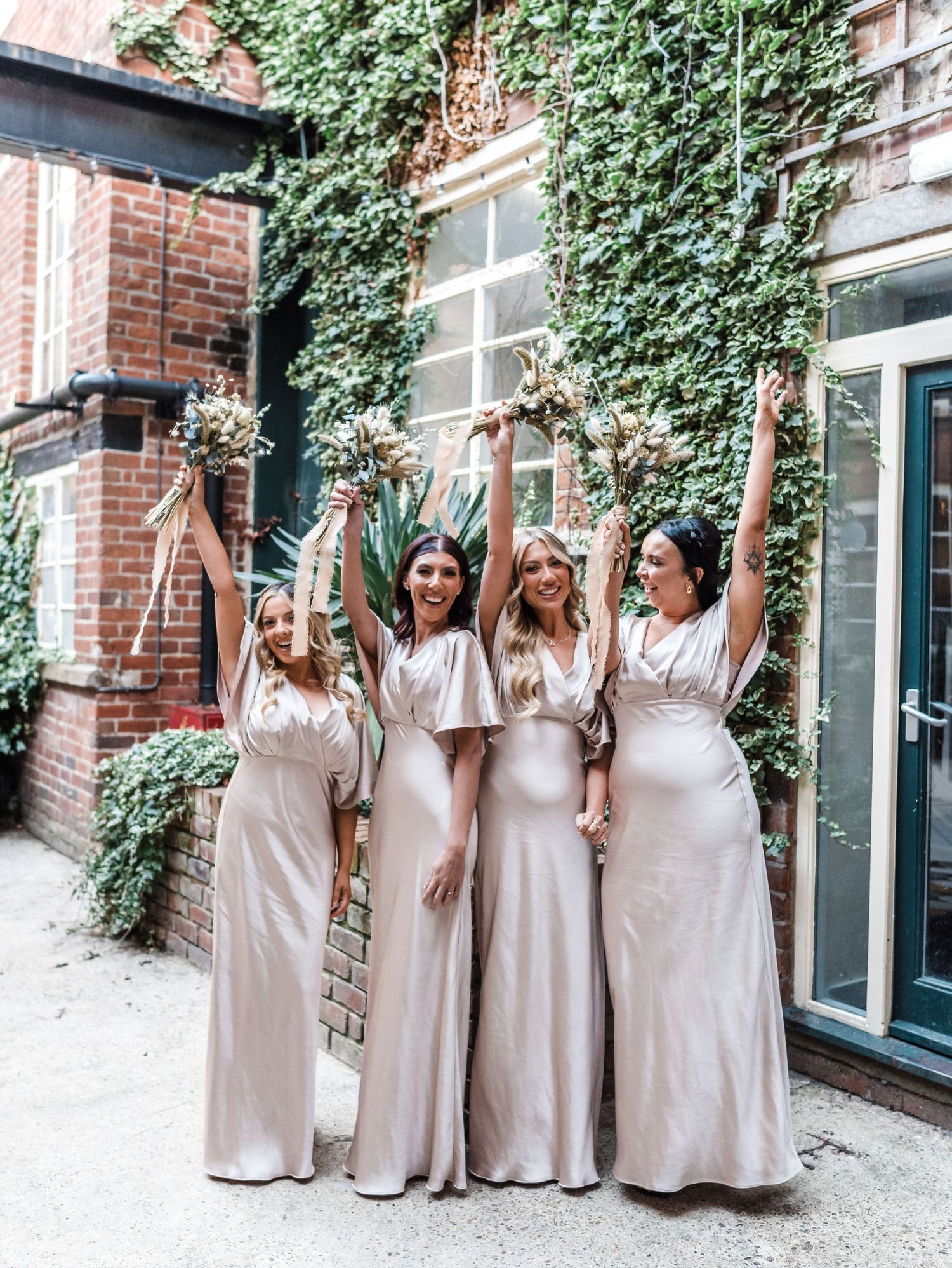 Four attendants in champagne-colored silk dresses pose against ivy-covered brick wall.