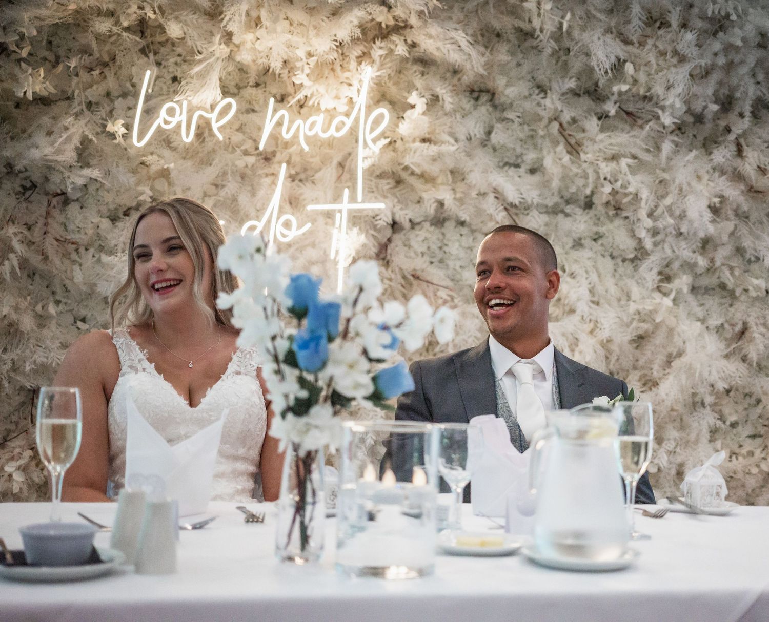 Wedding couple sits at reception table beneath glowing neon 'love you' sign with elegant floral decor.