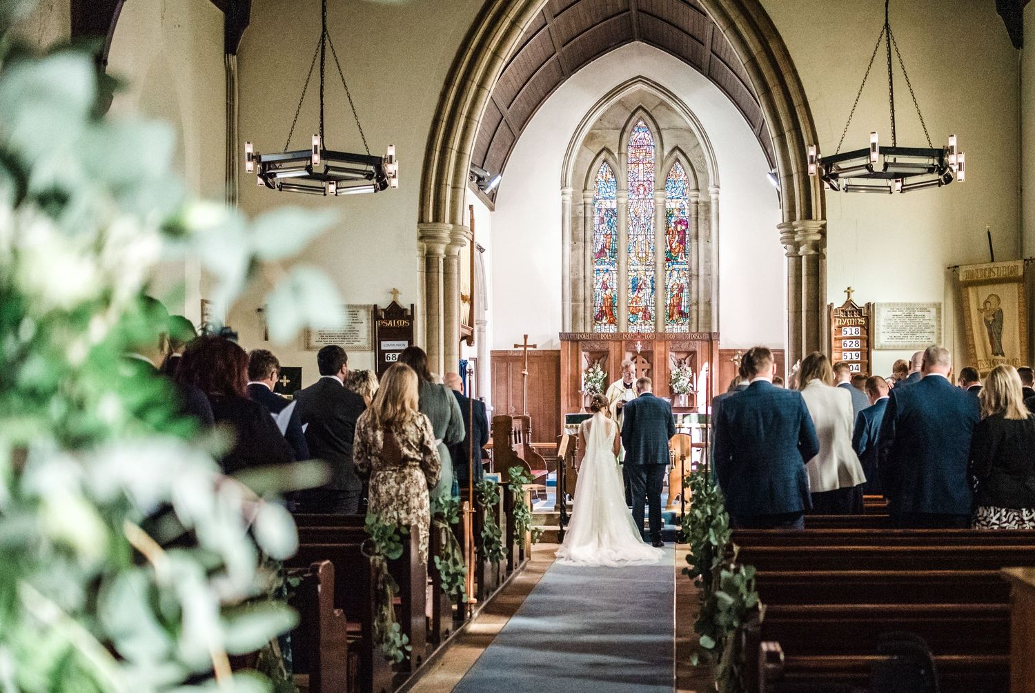 Elegant church wedding ceremony with stained glass windows and floral decorations along the aisle.
