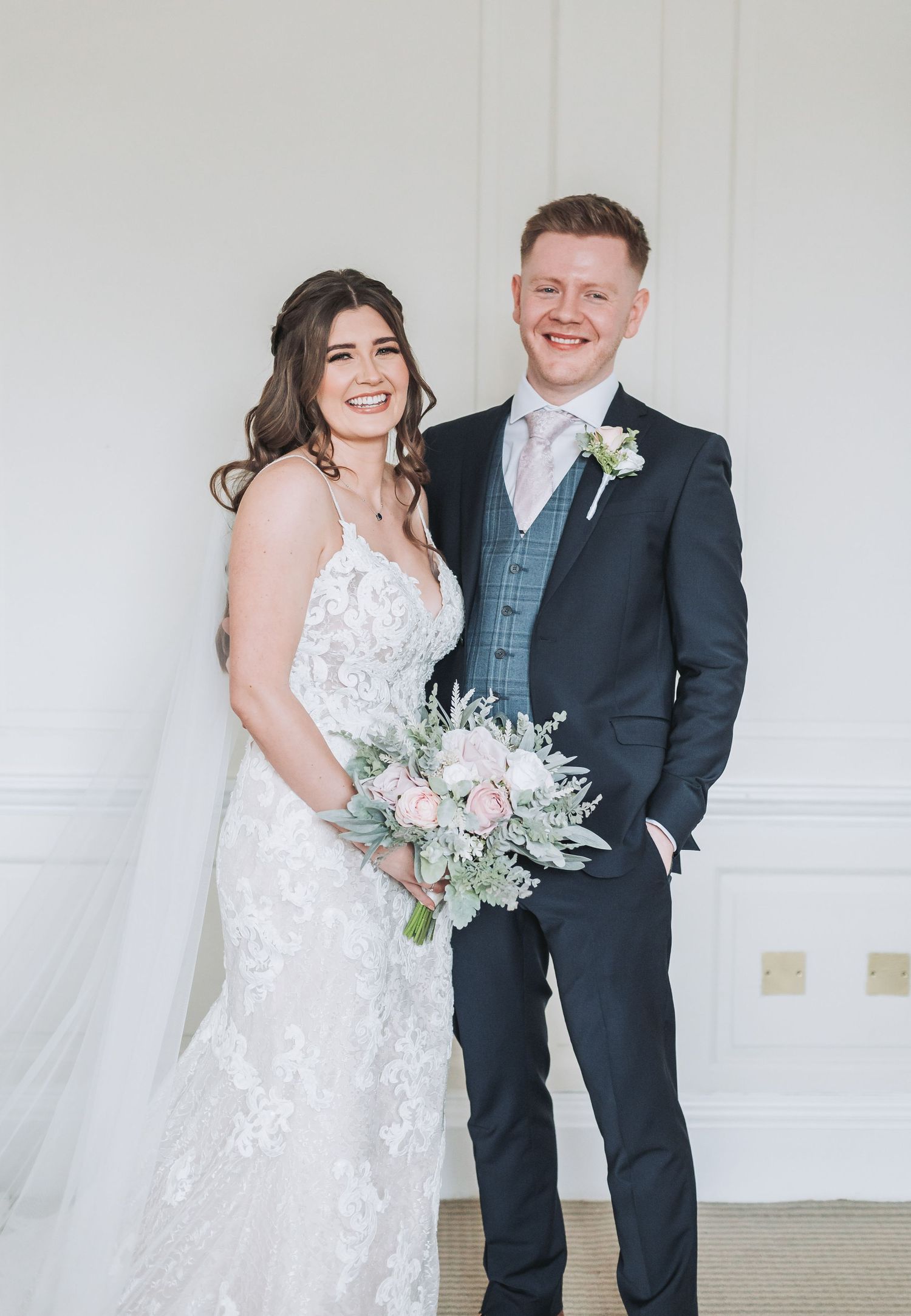 Wedding couple posing together against white wall with bride holding pastel bouquet.