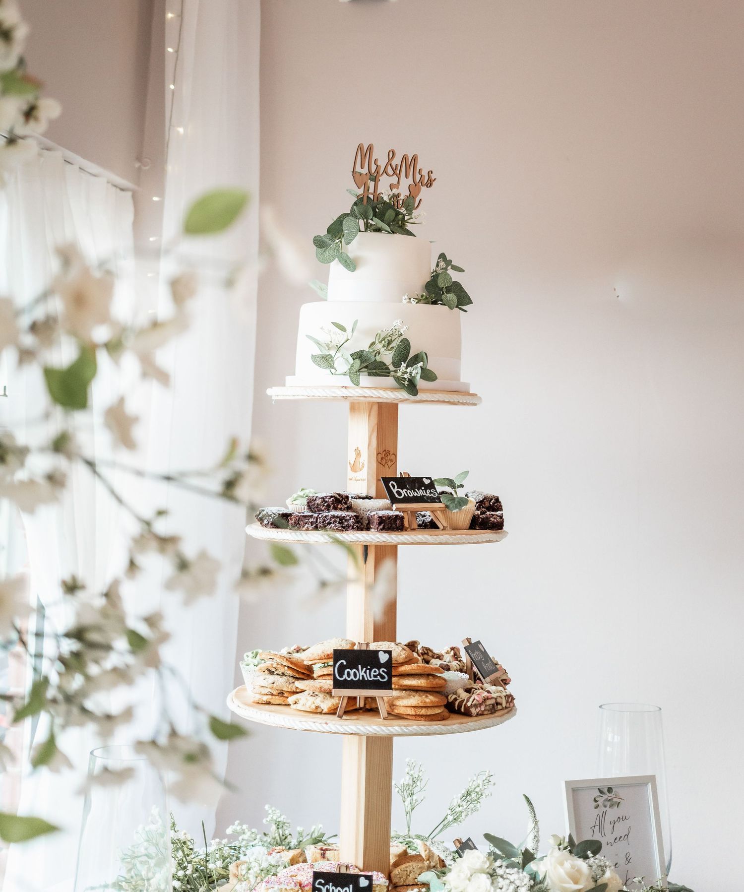 Multi-tiered white wedding cake display with greenery and desserts on wooden stand.
