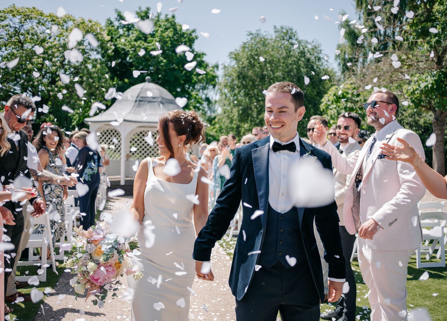 Wedding guests shower the newlyweds with confetti as they walk down an outdoor aisle on a sunny day.