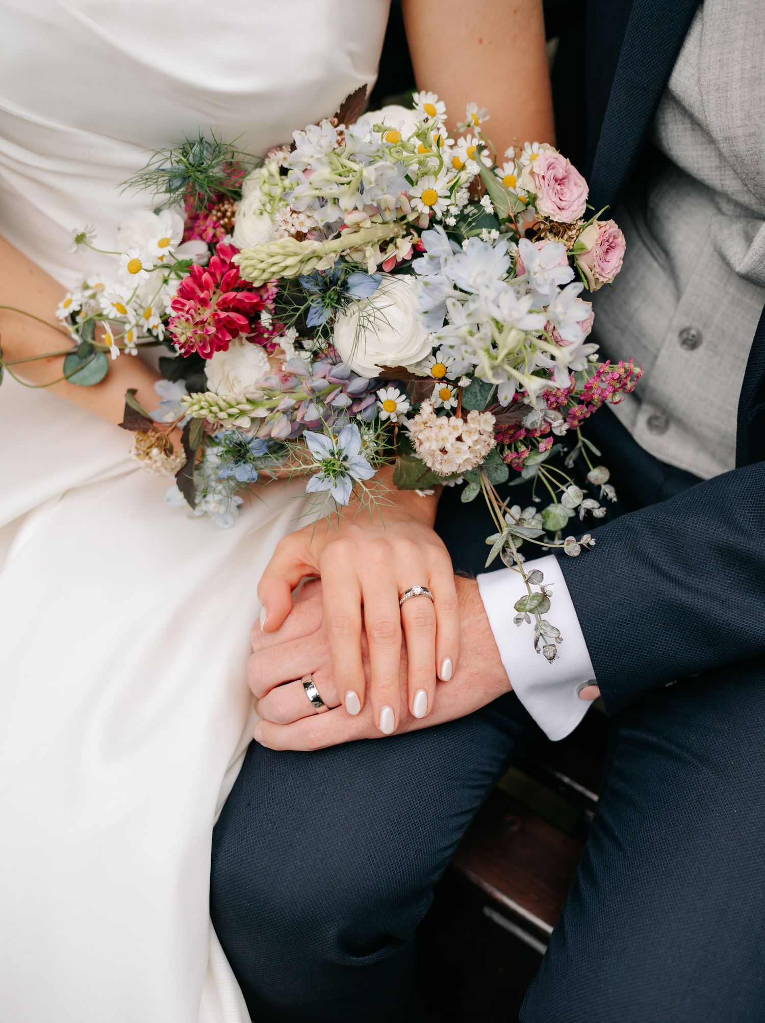 A stunning wildflower wedding bouquet rests on the couple's hands, showcasing their wedding rings.