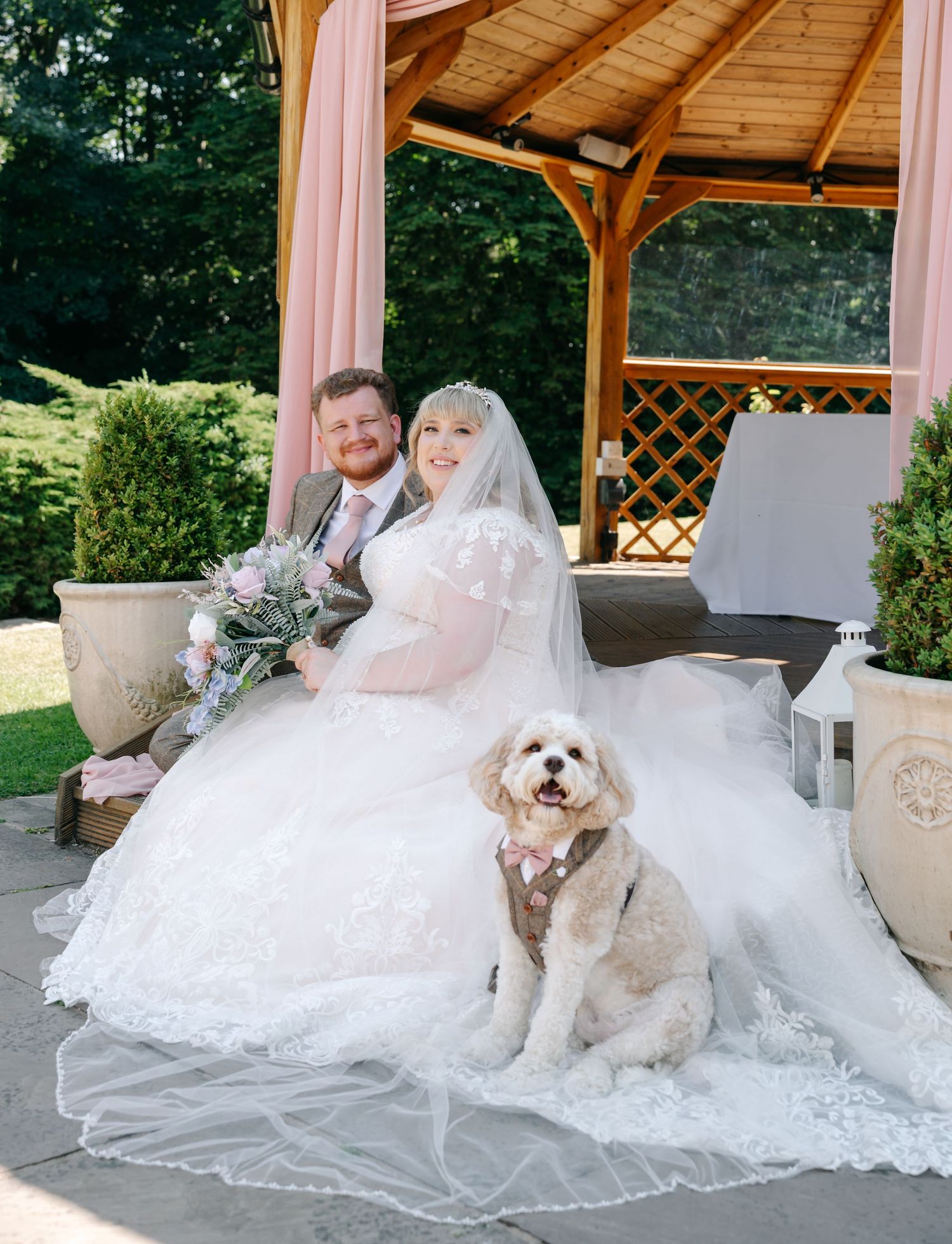 Bride and groom sitting together under a wooden gazebo with their dog in a bow tie on their wedding day.