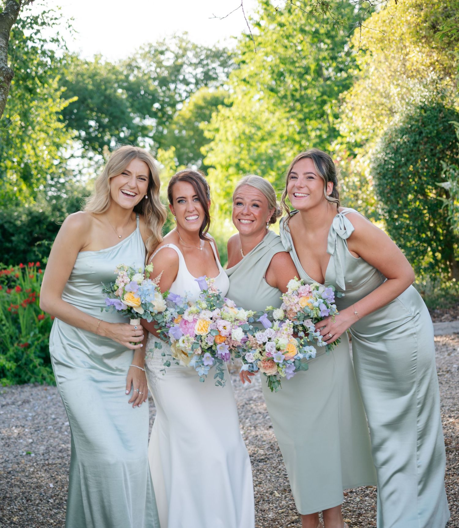 Bridesmaids in sage green dresses pose together with pastel bouquets in an outdoor garden setting.