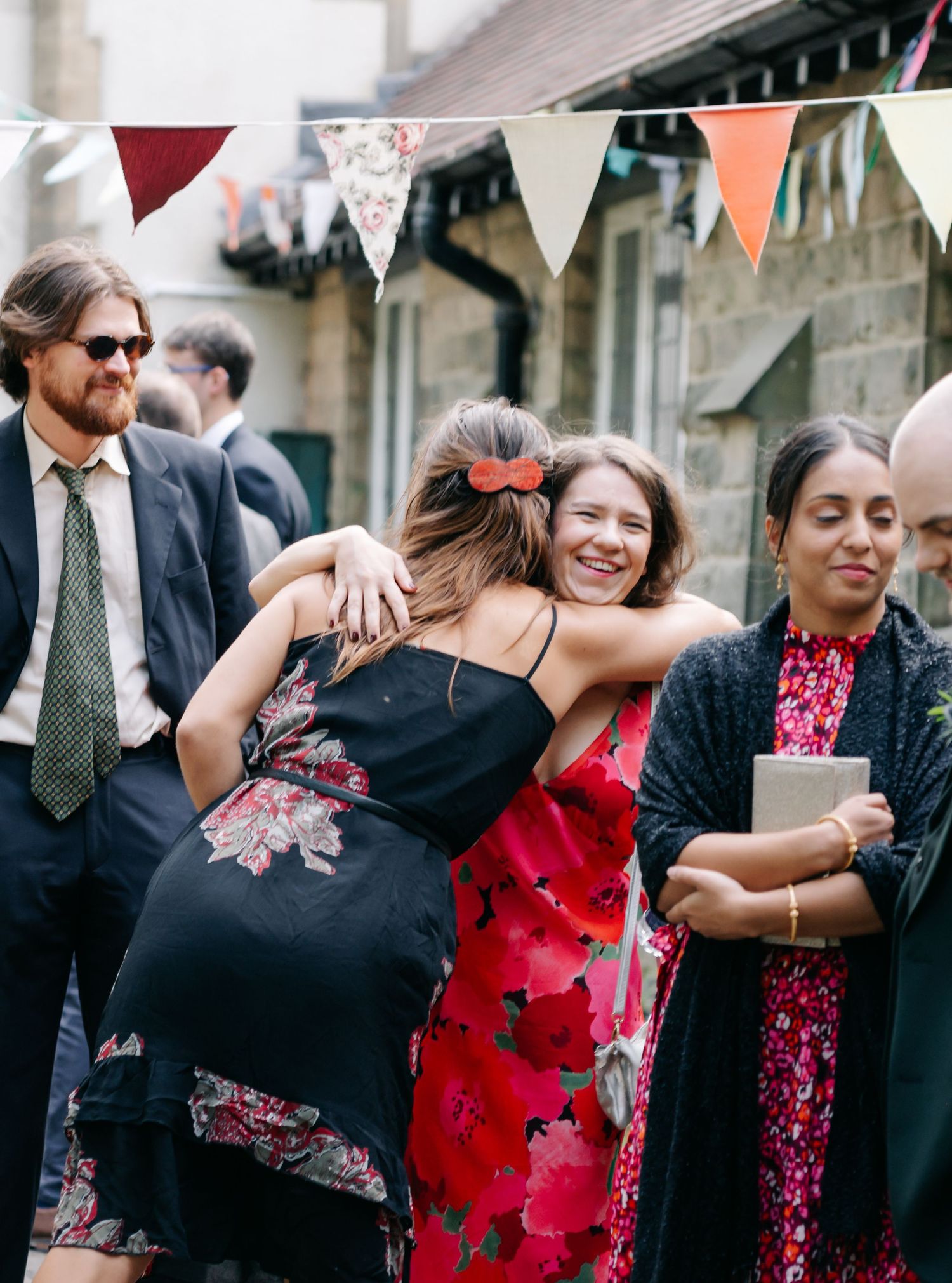 Wedding guests dance together in black and red attire at an outdoor celebration with decorative bunting overhead.