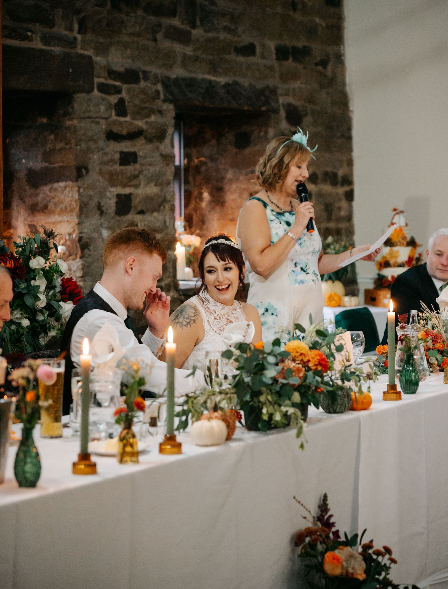 A decorated wedding reception table with orange flowers, greenery and candlelight against a rustic stone wall.