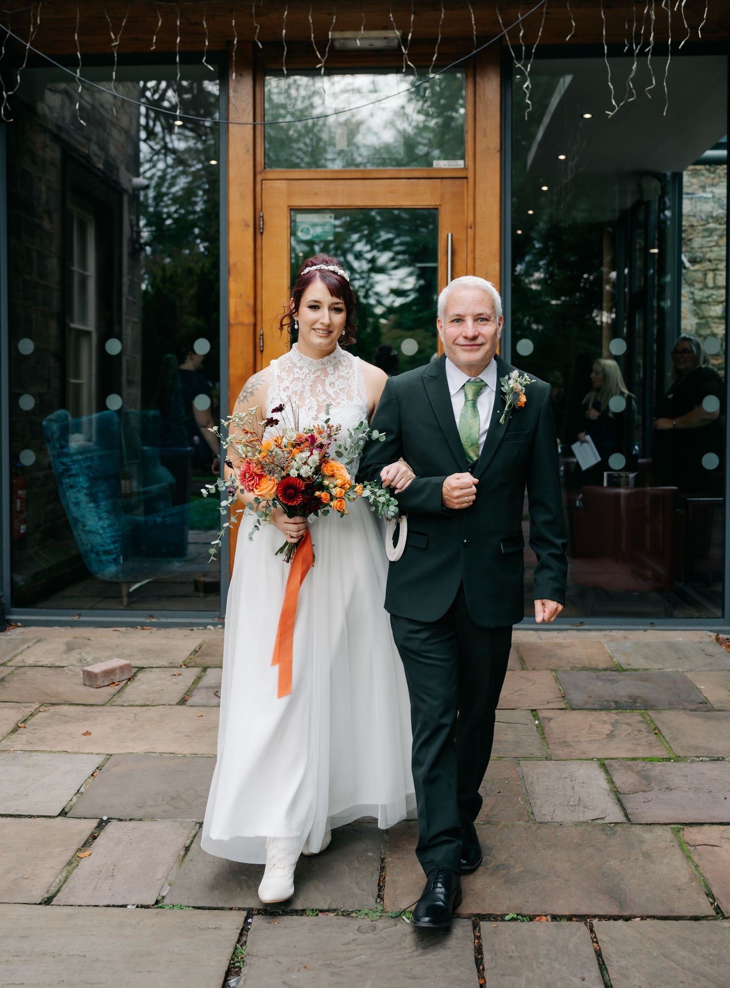 A wedding couple walks along a stone patio outside a venue holding colorful autumn bouquets.