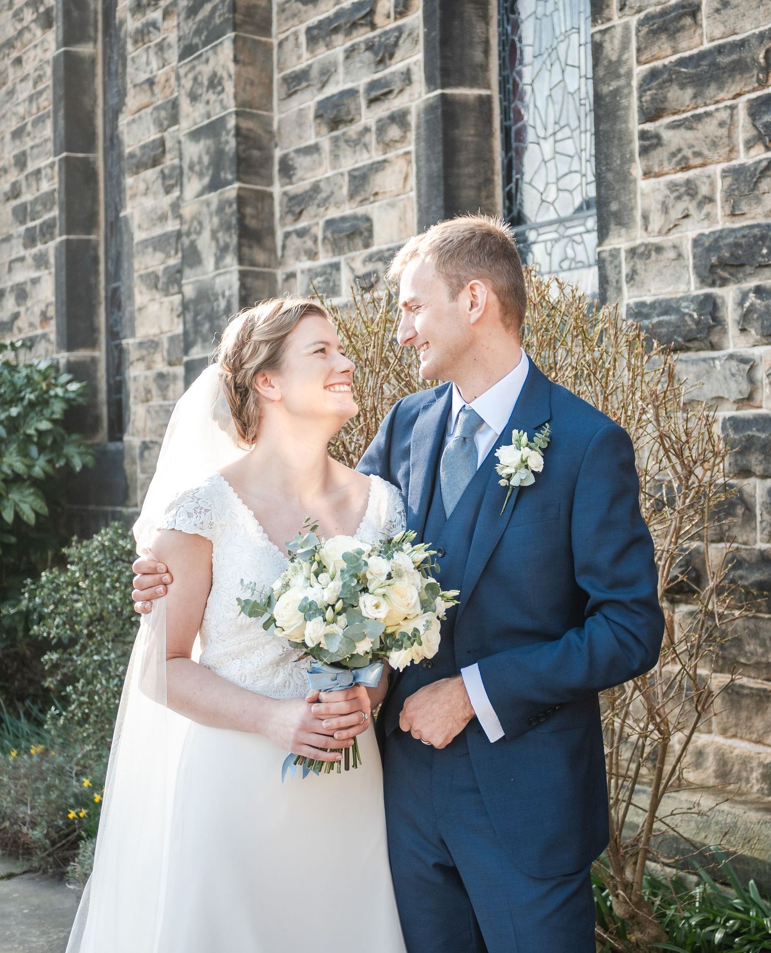 Newlyweds share a joyful moment against a stone wall backdrop with white floral bouquet.