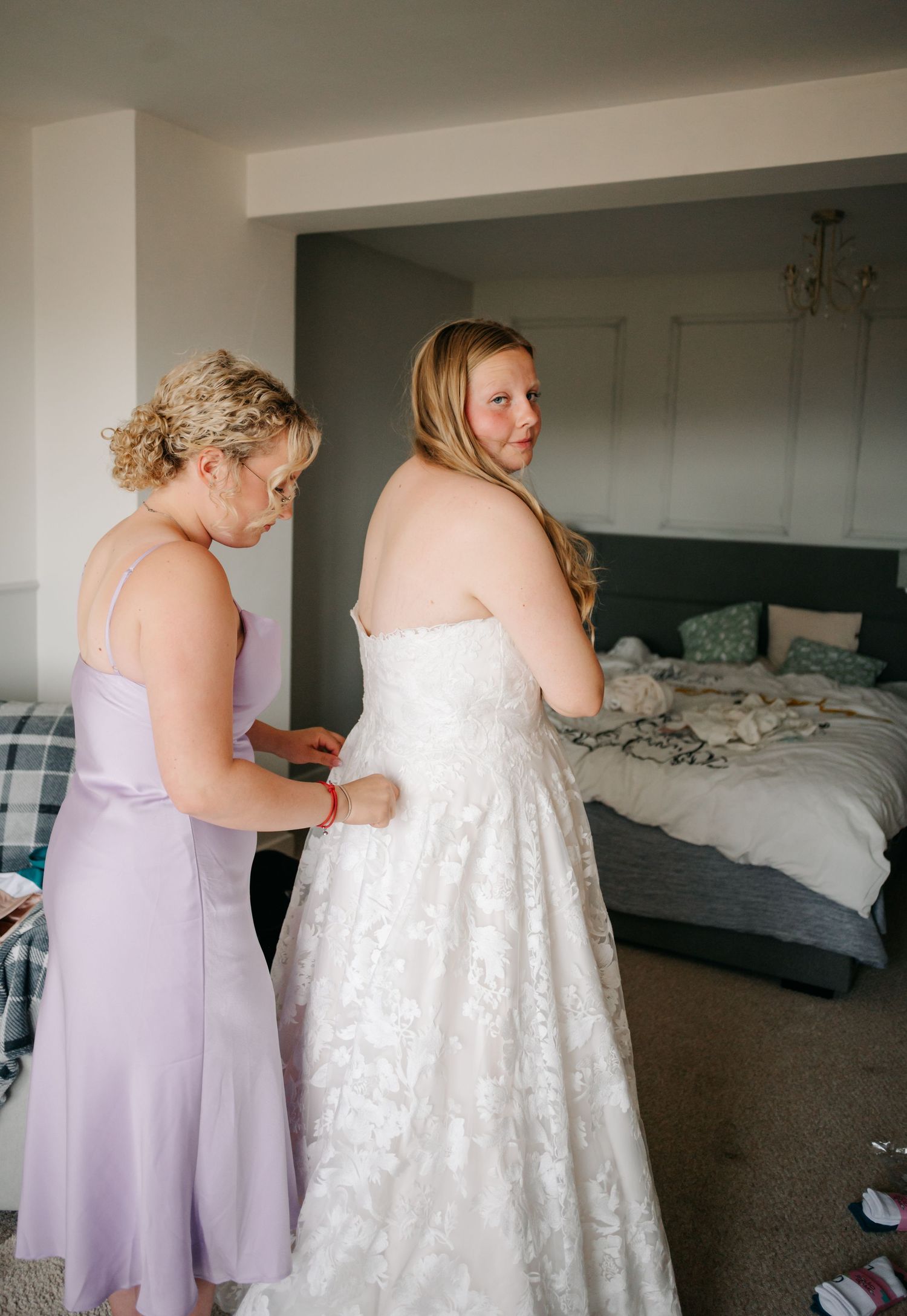 Bridesmaids in lavender dresses help button up a white lace wedding gown in a bedroom setting.