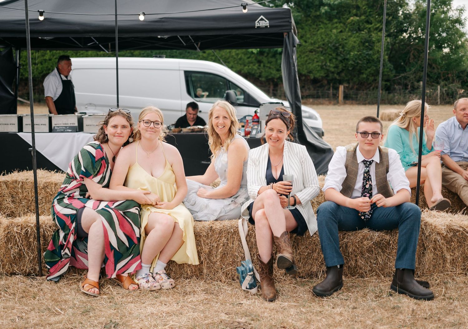 Wedding guests relax on hay bales during outdoor rustic farm reception.