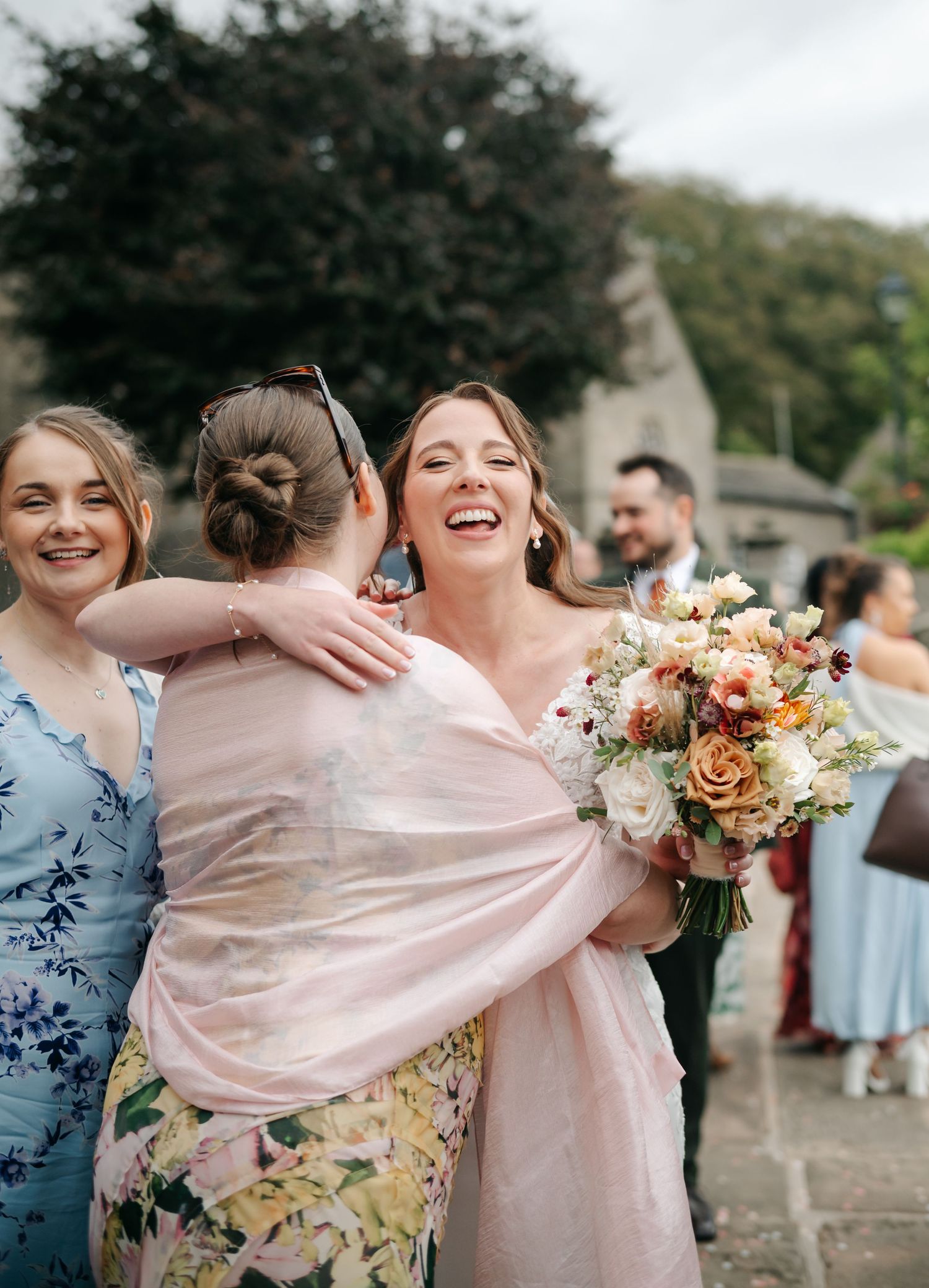 A laughing bride in a lace gown holds a floral bouquet while embracing a guest in a pink shawl outside a stone church.
