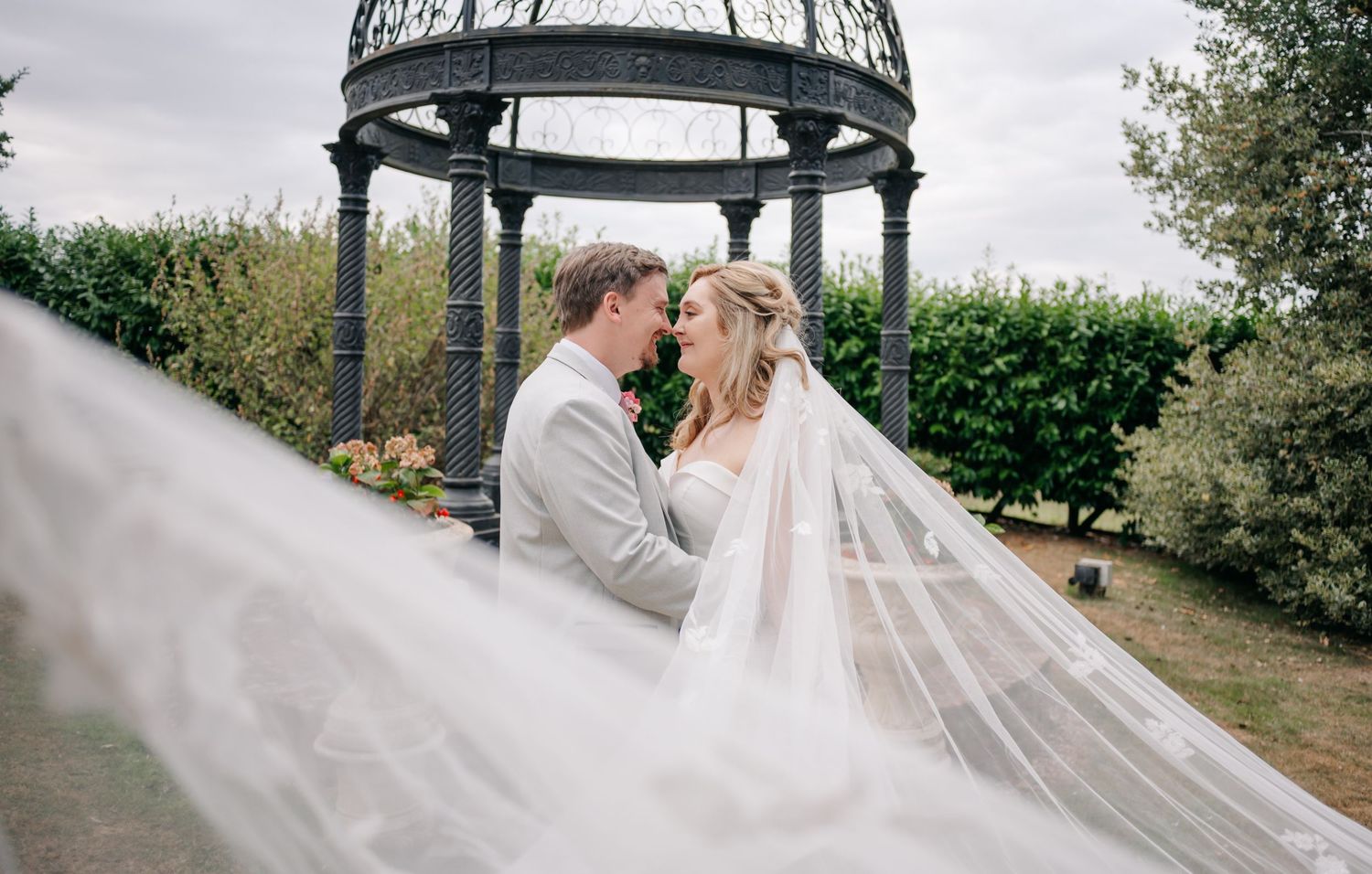 Wedding couple poses under an ornate gazebo with flowing veil in outdoor garden setting.