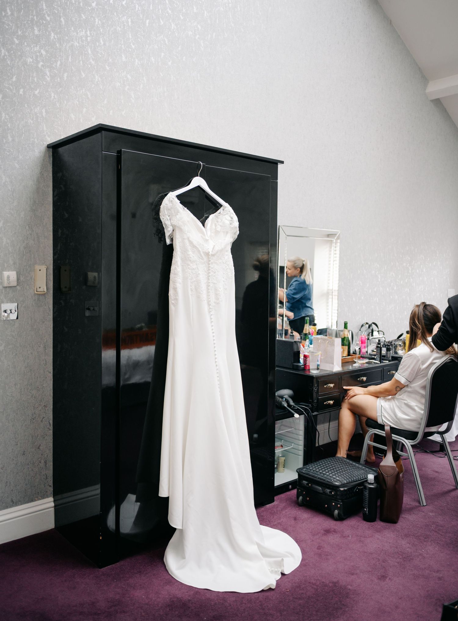 A white wedding dress hangs on a black wardrobe in a dressing room with a makeup station visible.