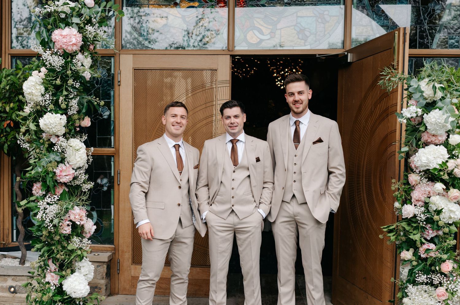 Wedding party in beige suits poses under elegant white floral arch decorations at venue entrance.