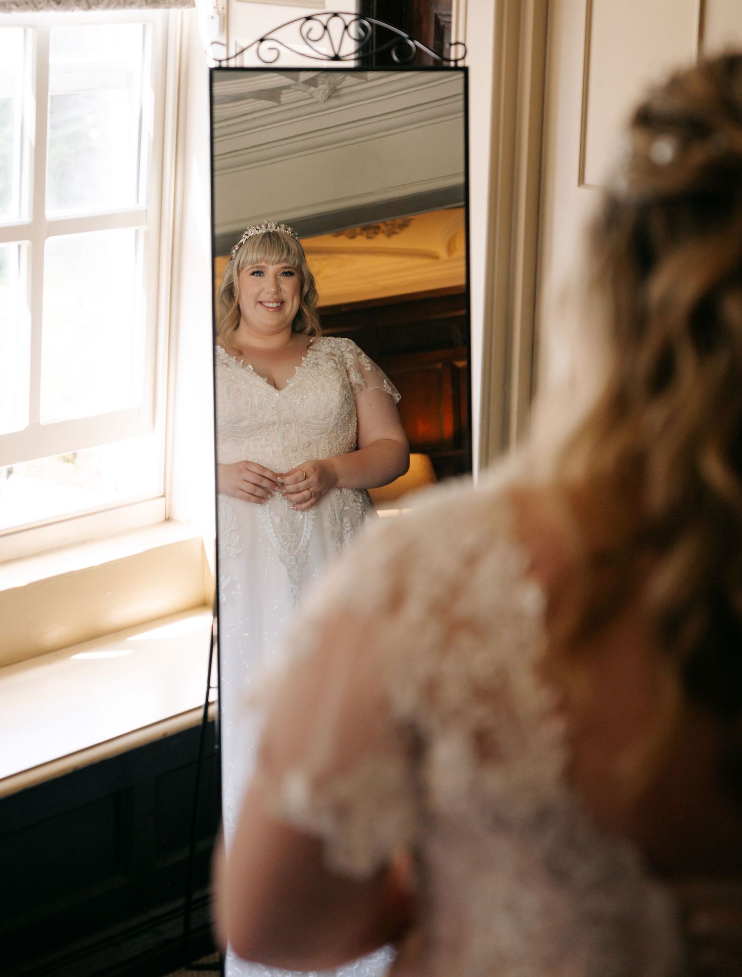 Bride in lace wedding gown and tiara admiring her reflection in a tall mirror before her wedding ceremony.
