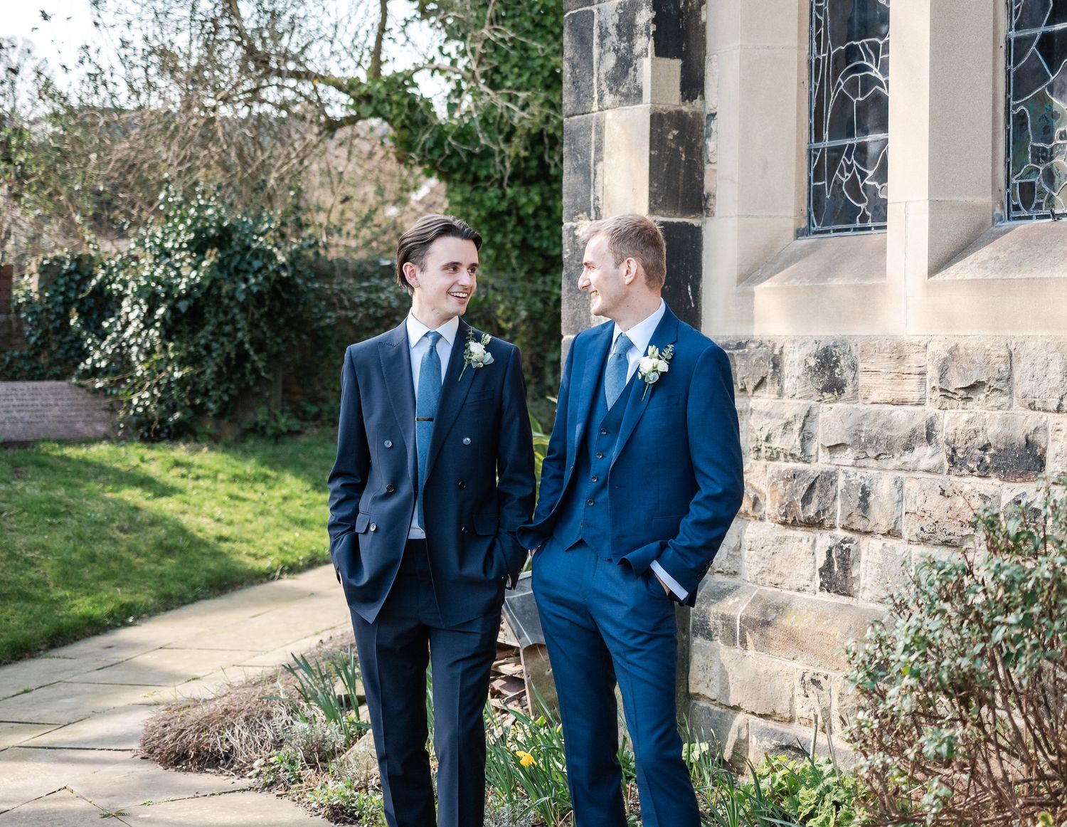 Two people in formal suits stand together outside a stone building in conversation.