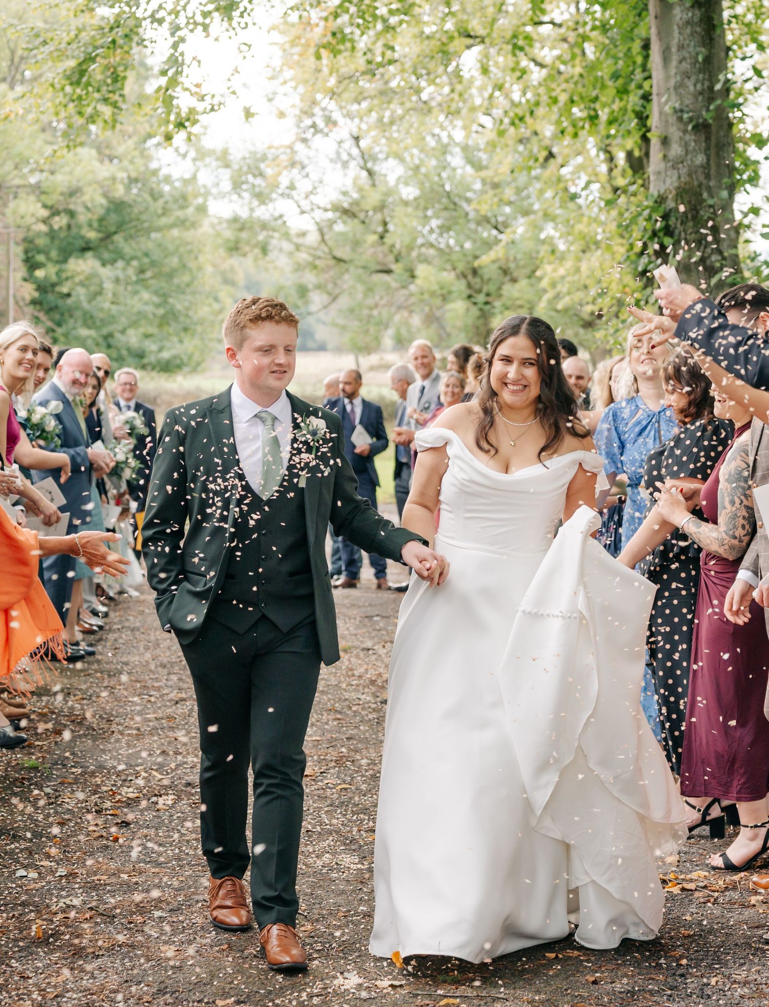 Wedding guests shower newlyweds with confetti on a tree-lined path during autumn celebration.