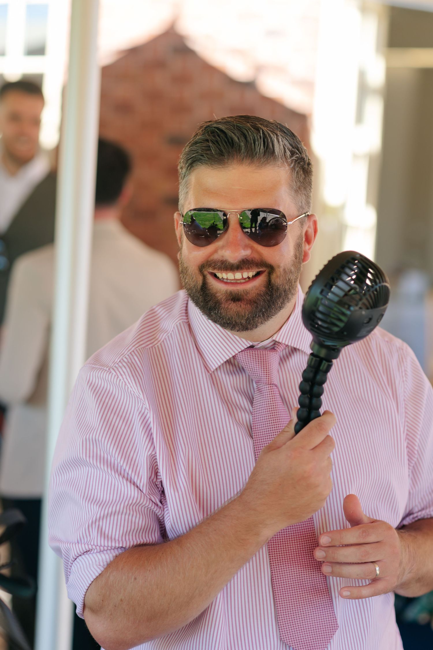 Wedding guest in pink striped shirt and sunglasses holds a microphone while smiling and laughing at an outdoor event.