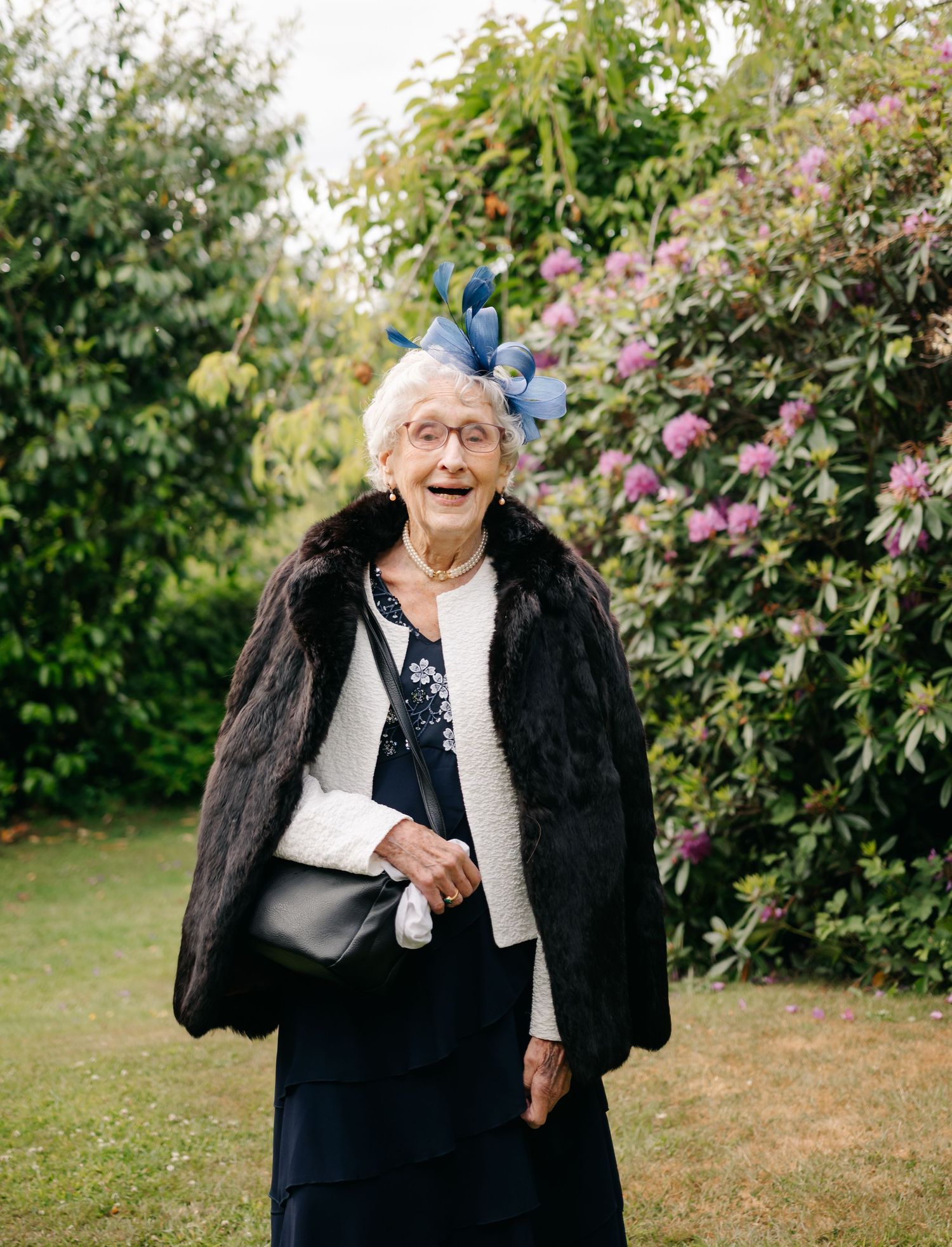 Joyful elderly guest wearing a fur coat and blue butterfly fascinator in a garden setting.