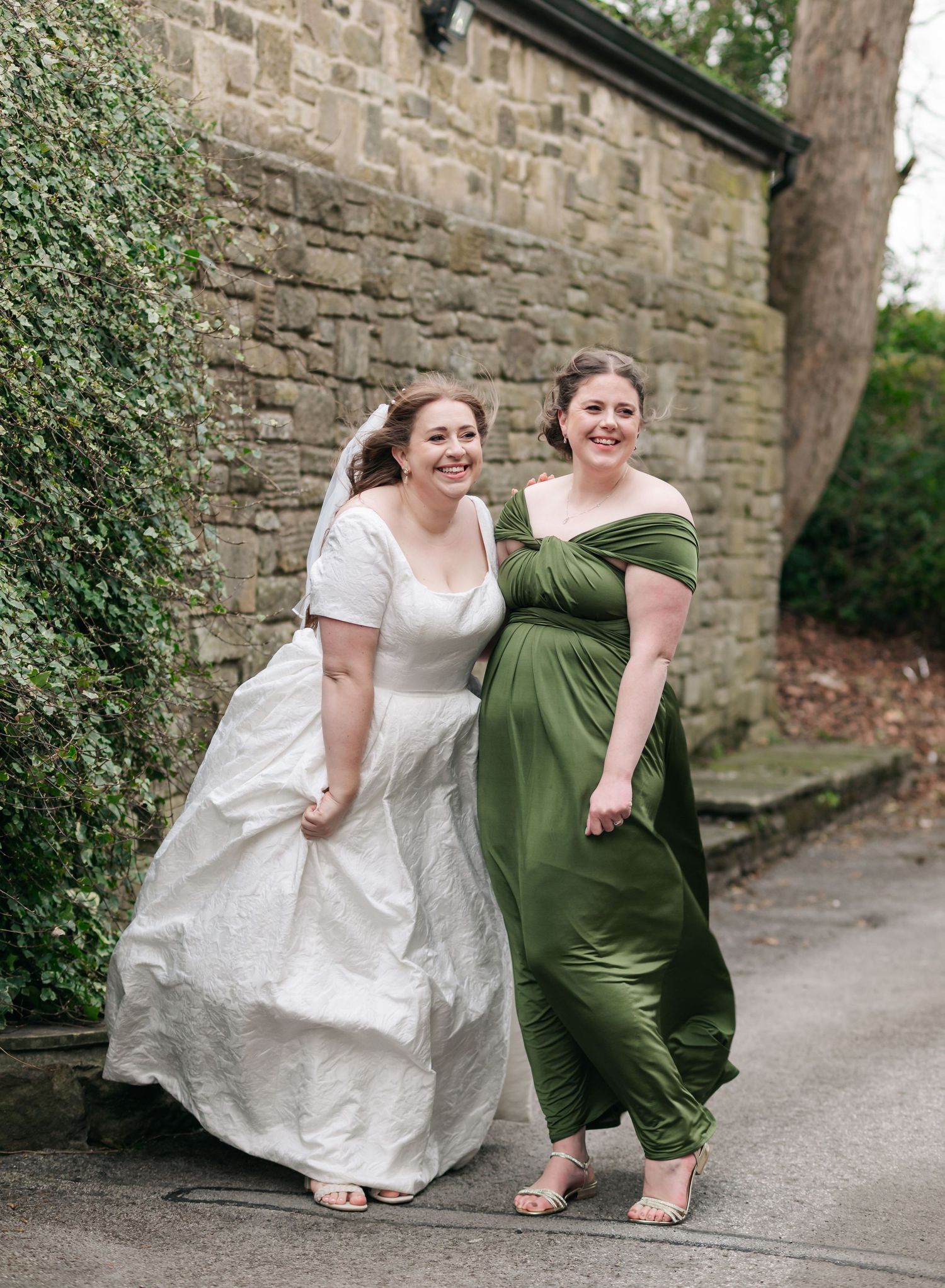 Two people in white and olive green dresses share a playful moment on a stone path by a rustic wall.
